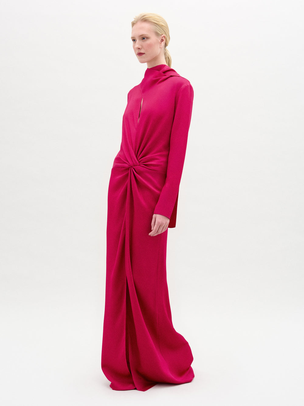 A person stands against a plain white background wearing the Sue Dress by SVT DESIGNS—a long-sleeved, bright pink maxi dress crafted from luxurious Italian fabric, featuring a high neckline and a gathered knot detail at the waist.