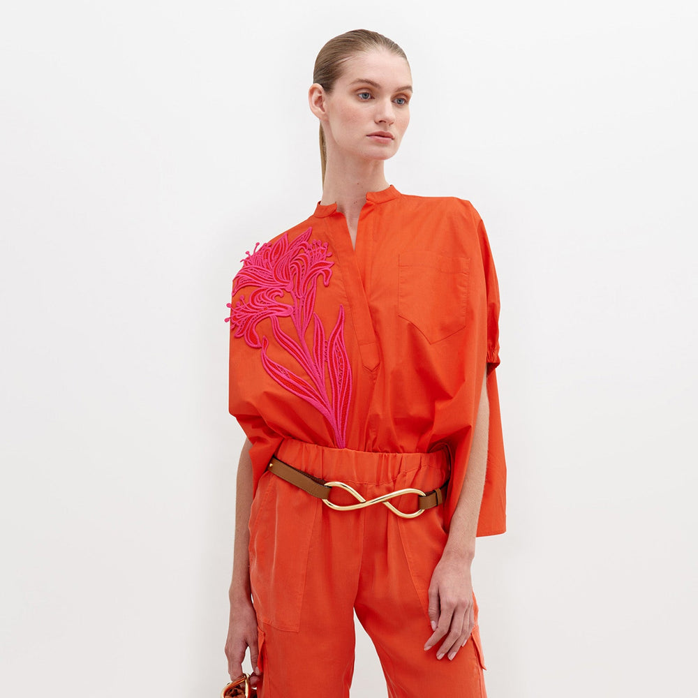 A woman poses against a white background in the Susanne Blouse by SVT DESIGNS—a voluminous, 100% cotton orange top with a high collar, short sleeves, a large pink floral motif on one side, and a tan belt with a gold wavy buckle.