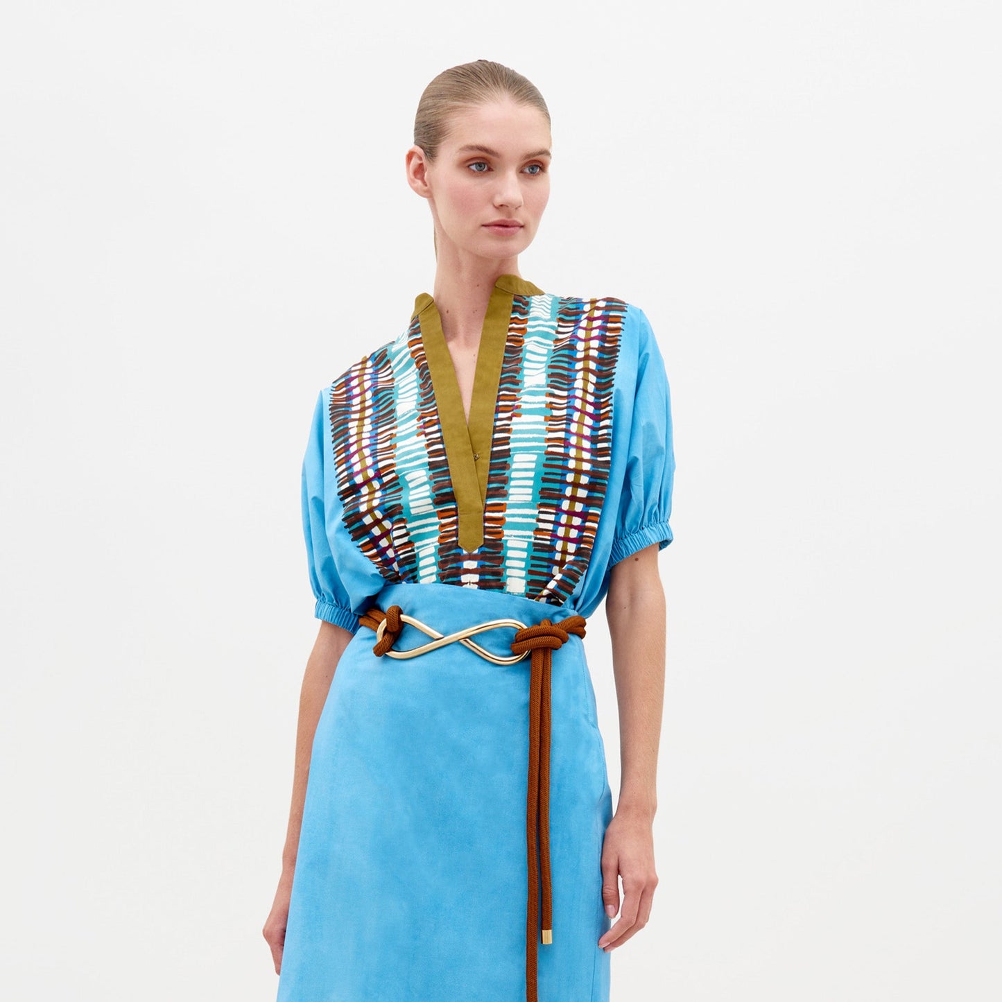 A woman stands against a white background wearing the Susanne Blouse by SVT DESIGNS—a blue cotton top featuring short sleeves, an abstract print V-neck bodice, and a brown rope belt with gold accent. Her hair is neatly pulled back.