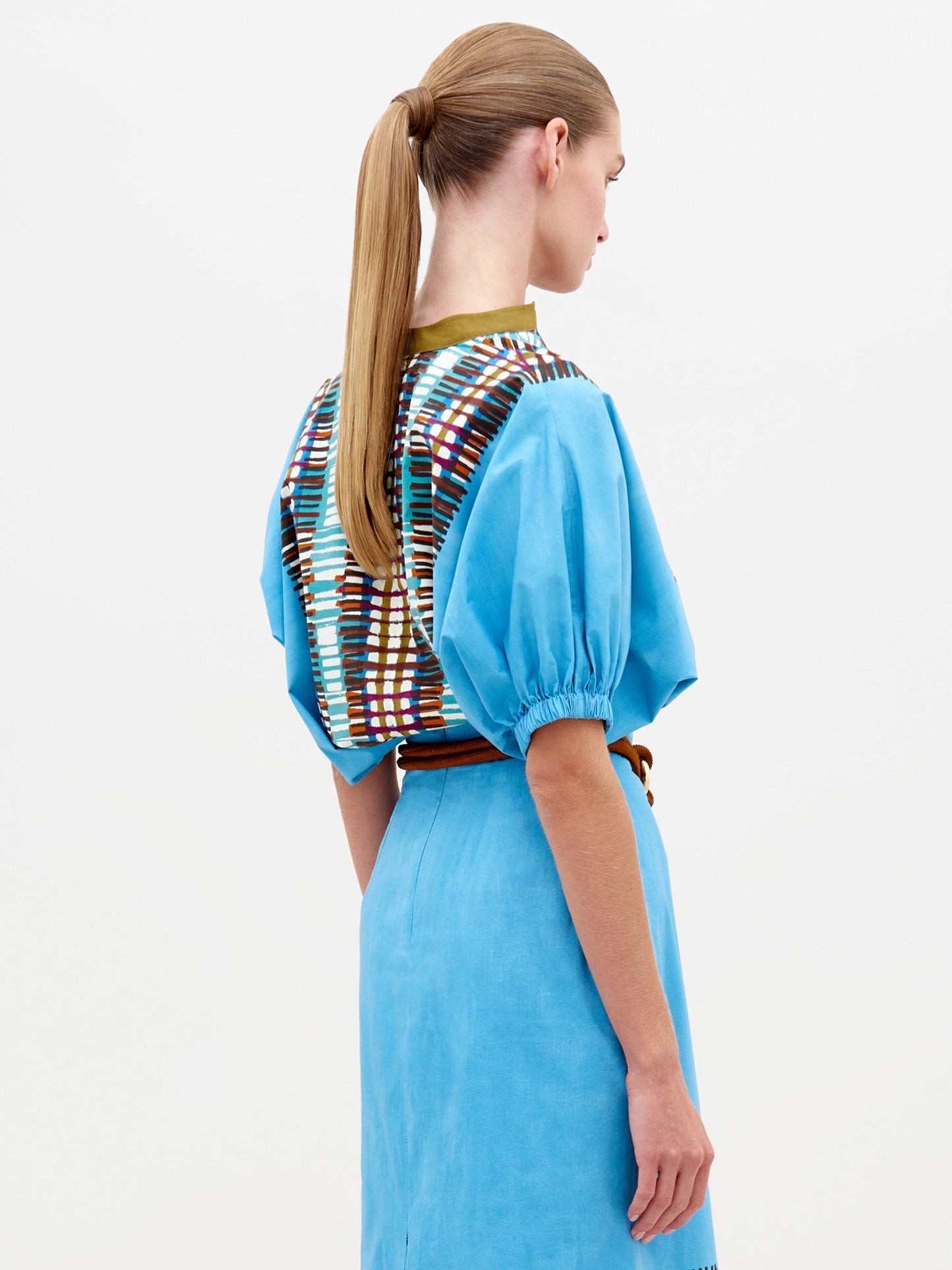 A woman wears the Susanne Blouse in Aqua Olive Plaid, featuring a multi-colored patterned V-neck and a knotted brown belt, as she stands against a plain white background. This elegant outfit is available for pre-order now and will ship by February 15th, 2025.