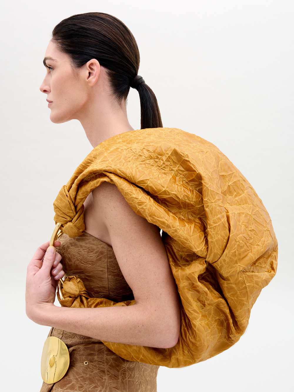 A person with dark hair tied back wears a textured, golden-brown outfit and carries the Sofia Bag Maxi by SVT DESIGNS, an Italian fabric bag with a matching crumpled texture, over one shoulder against a plain white background.