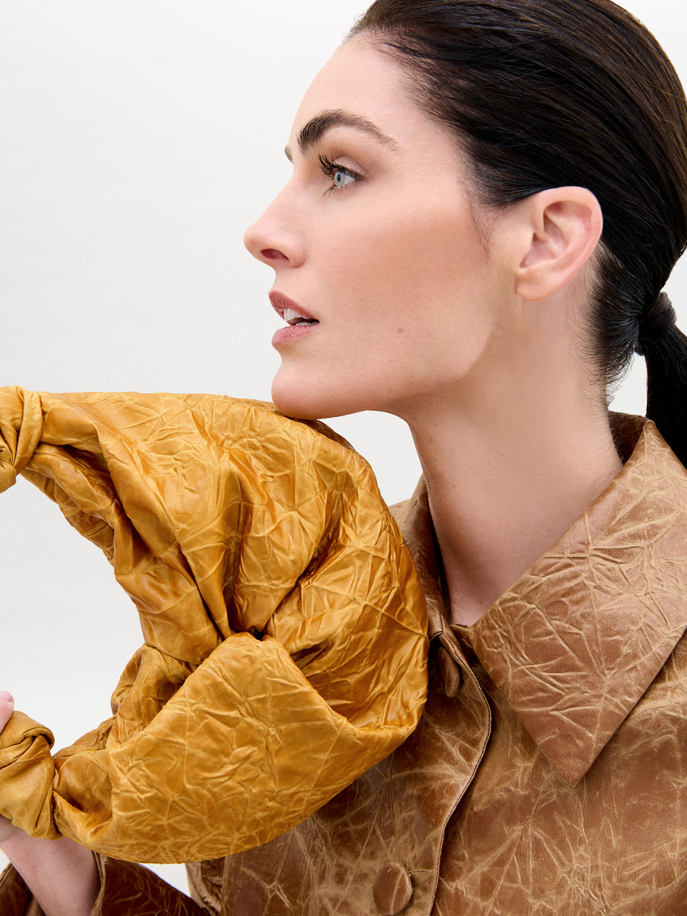 A person with dark hair tied back holds the Sofia Bag Small from SVT DESIGNS, featuring a textured mustard color and an S-shaped gold-toned handle, near their face. They wear a matching textured coat and look to the side against a plain background.