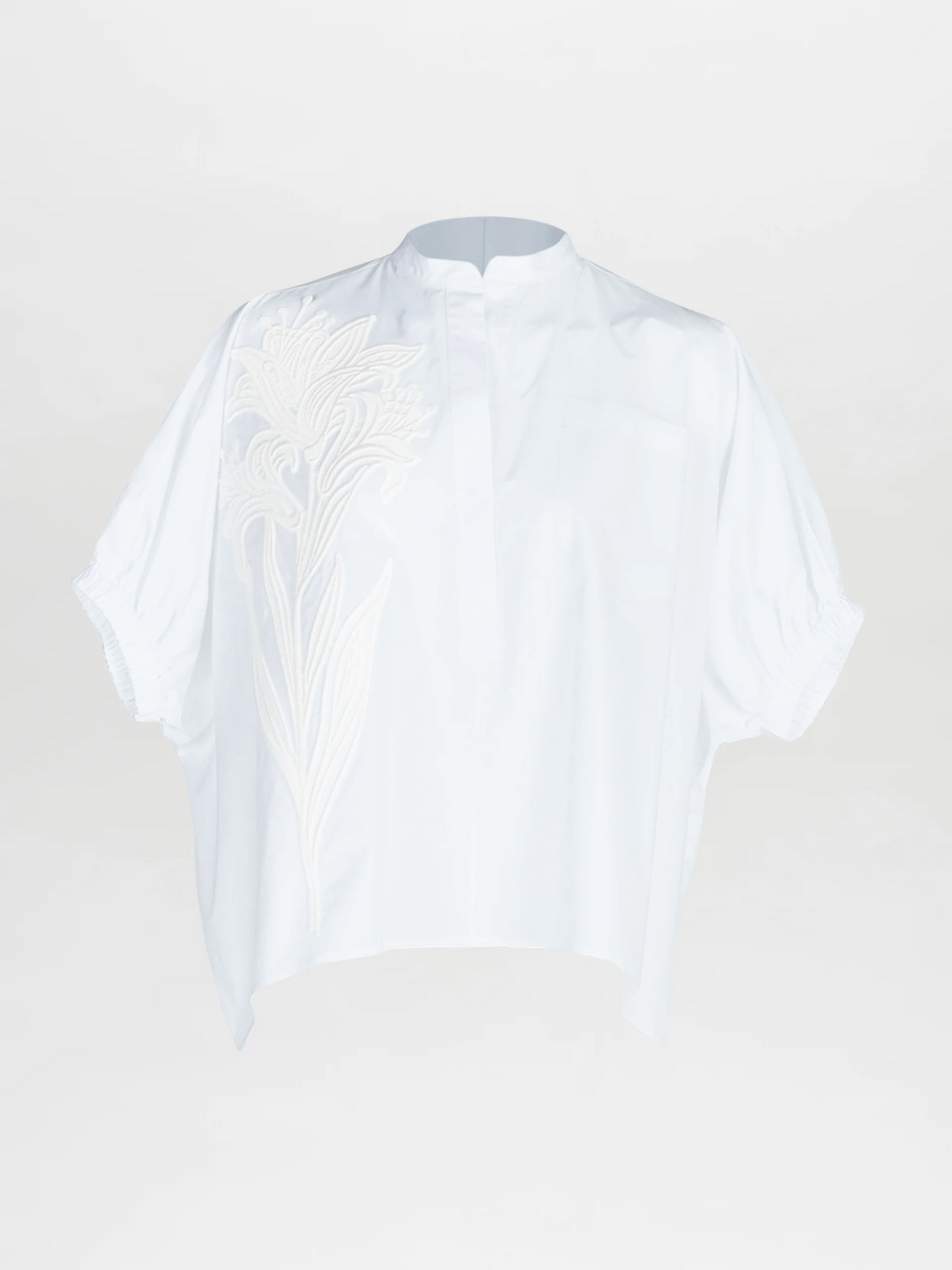 The Susanne Blouse is a loose-fitting, short-sleeved white top featuring a stand collar, subtle button placket, and oversized floral embroidery on the left shoulder and chest.