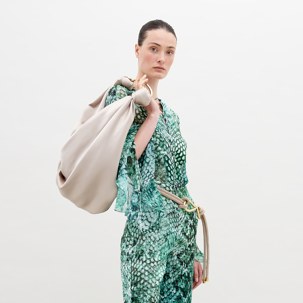 A person in a stylish green, abstract print outfit from SVT DESIGNS wears the Thea Blouse against a plain background. They carry a large, beige soft bag with knotted handles over their shoulder, and their hair is slicked back.
