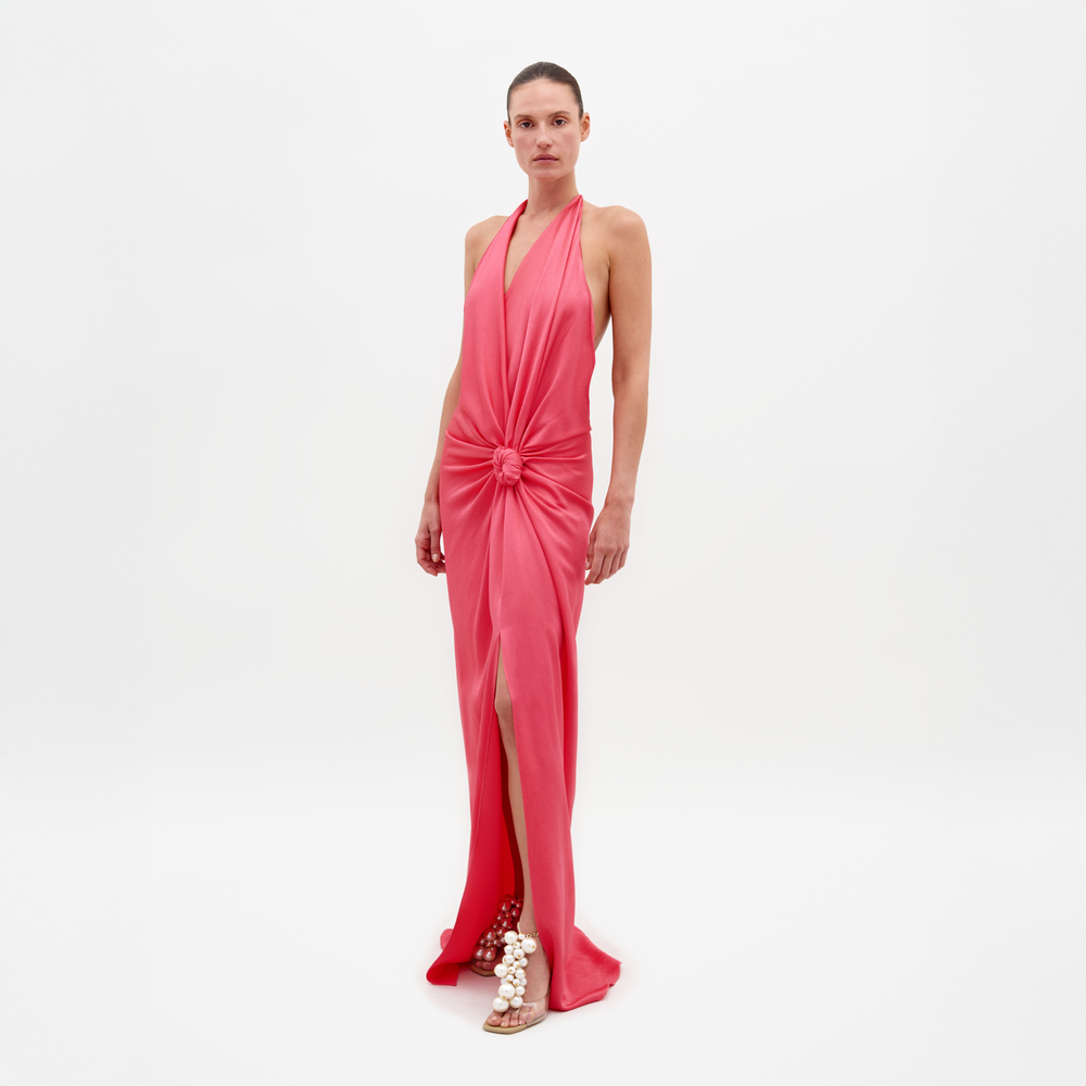 A person stands against a plain white background wearing the SVT DESIGNS Torgiano Dress—a sleeveless, floor-length coral pink maxi with a halter neckline, front knot detail, and high slit—paired with pearl-embellished flat sandals.