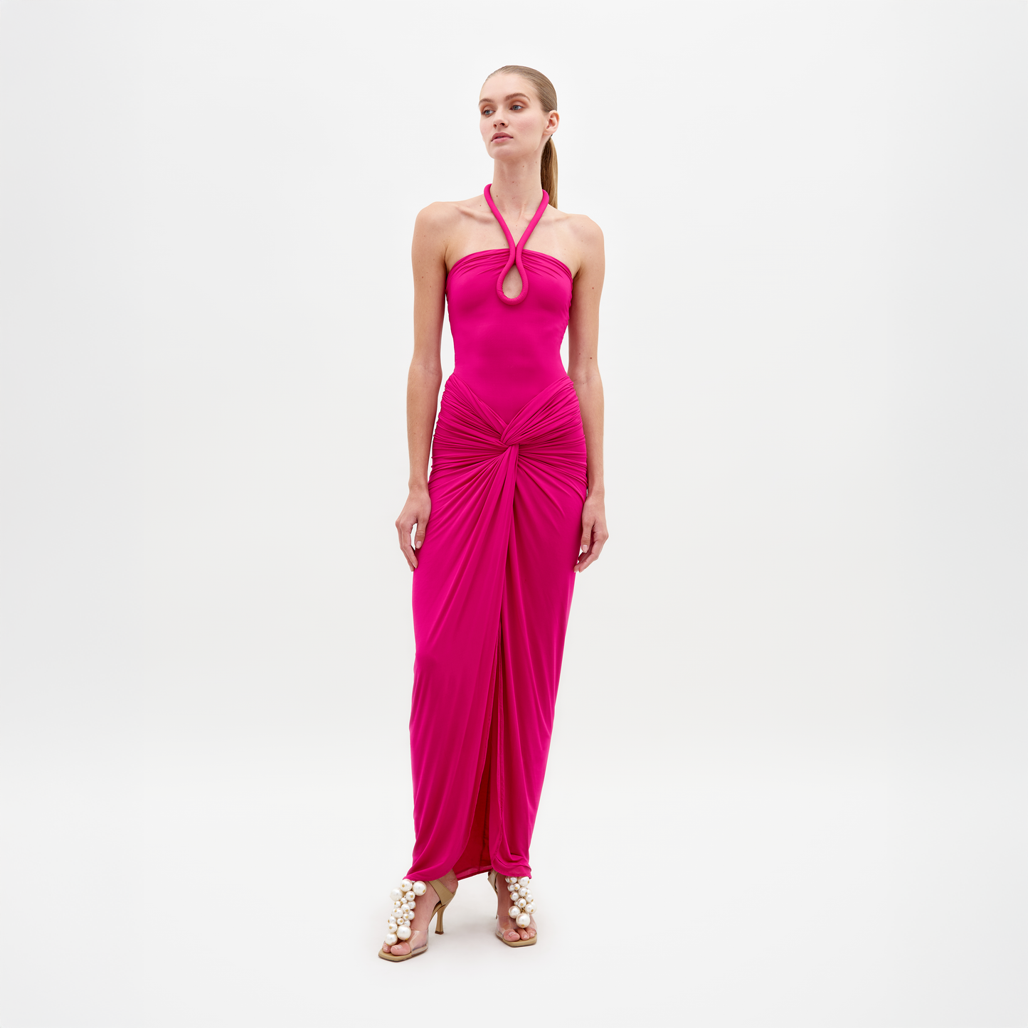 A woman poses against a white background wearing the SVT DESIGNS Tori Dress, a bright pink halter-neck maxi in luxurious Italian fabric with gathered draping at the waist and hem, styled with heeled sandals embellished with large pearls.