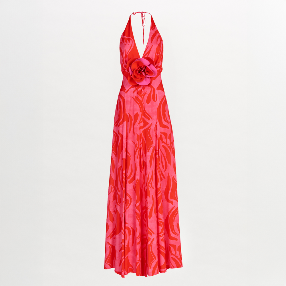 The Silvia Tcherassi Tawny Dress is a sleeveless, floor-length gown in red and pink with a halter neckline, open back, abstract swirl pattern, large waist flower accent, subtle sheen, and flowing silhouette for an elegant look.
