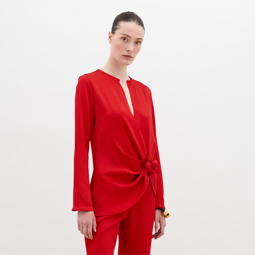 A person poses elegantly in the SVT DESIGNS Tayra Blouse, a long-sleeved hammered satin red top with a waist knot detail, paired with matching pants. Made in Portugal, the outfit creates a striking contrast against the plain white background.