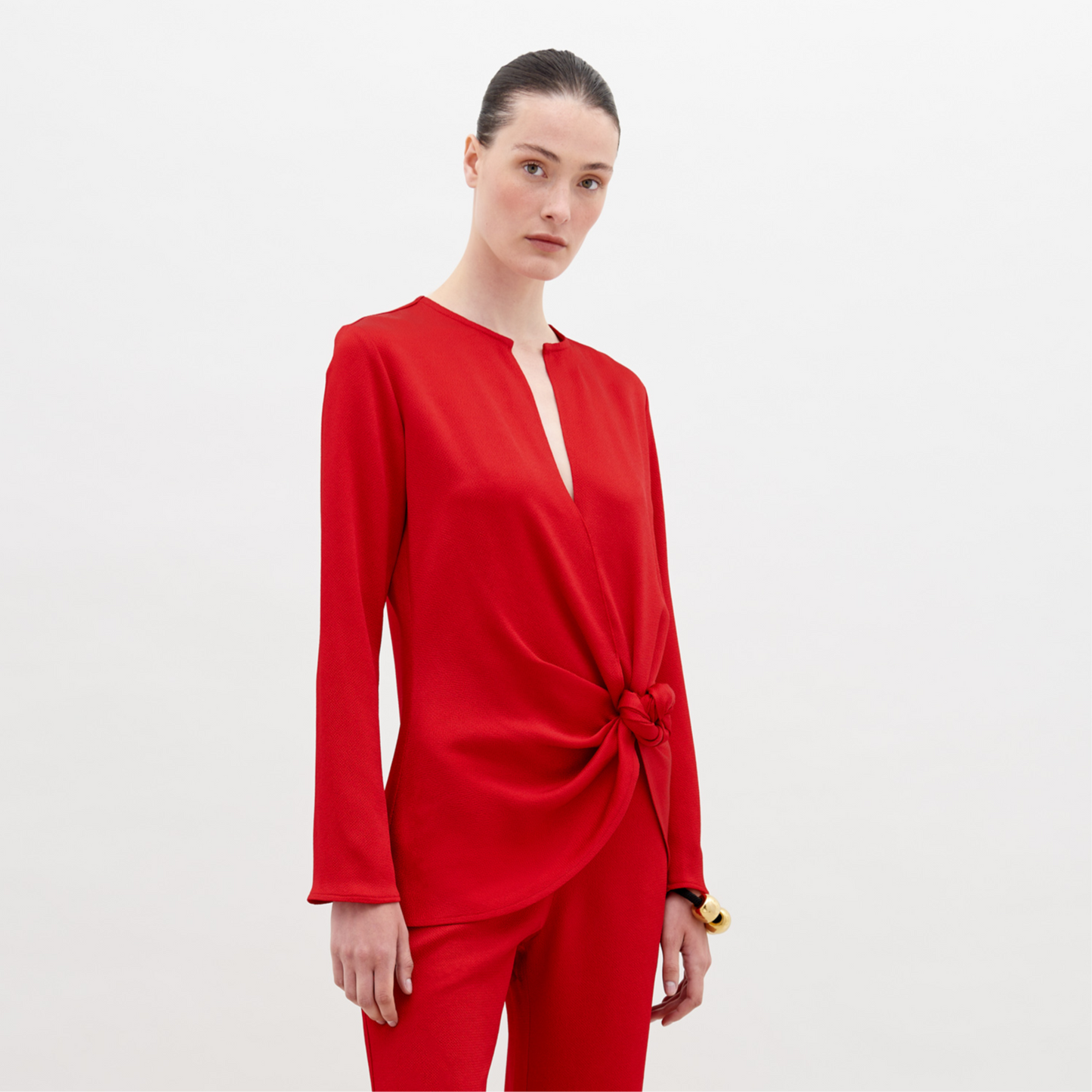 A person poses elegantly in the SVT DESIGNS Tayra Blouse, a long-sleeved hammered satin red top with a waist knot detail, paired with matching pants. Made in Portugal, the outfit creates a striking contrast against the plain white background.