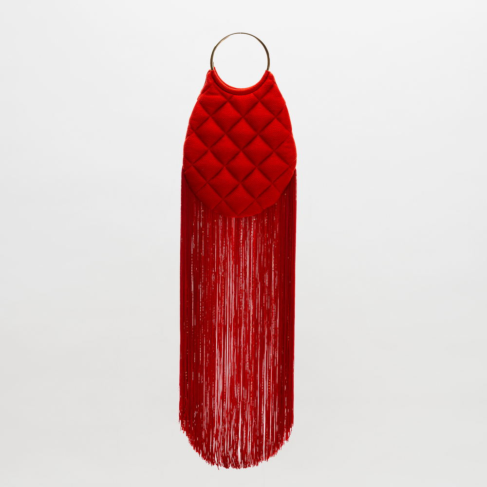 The SVT DESIGNS Toblach Bag is a red handbag with a quilted oval top, metallic top handles, and long fringe at the bottom, shown against a plain white background.