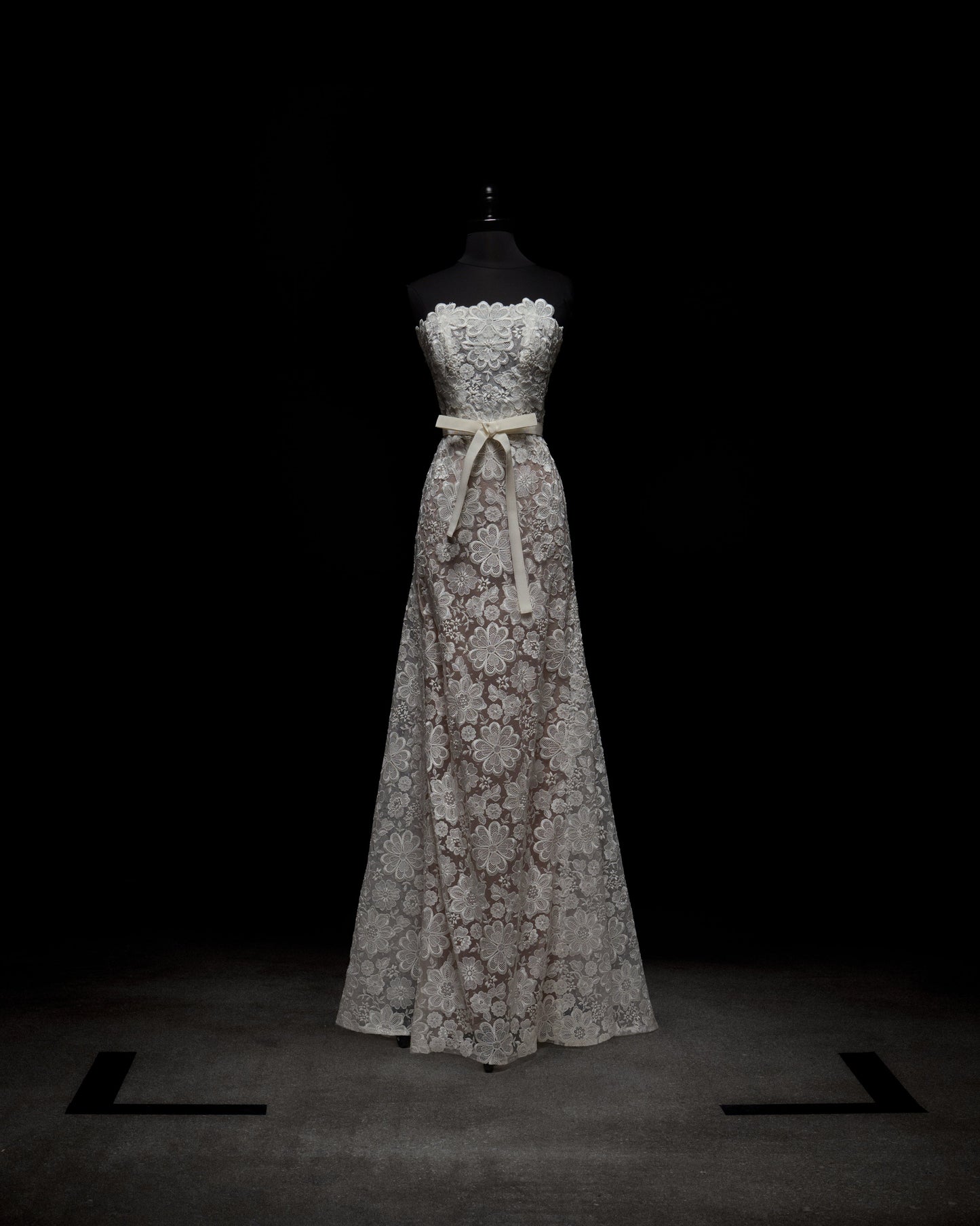 The Valencia Dress by SVT DESIGNS is a strapless, floor-length gown displayed on a mannequin against a dark backdrop. It showcases intricate white lace over light fabric and is accented with a beige ribbon at the waist.