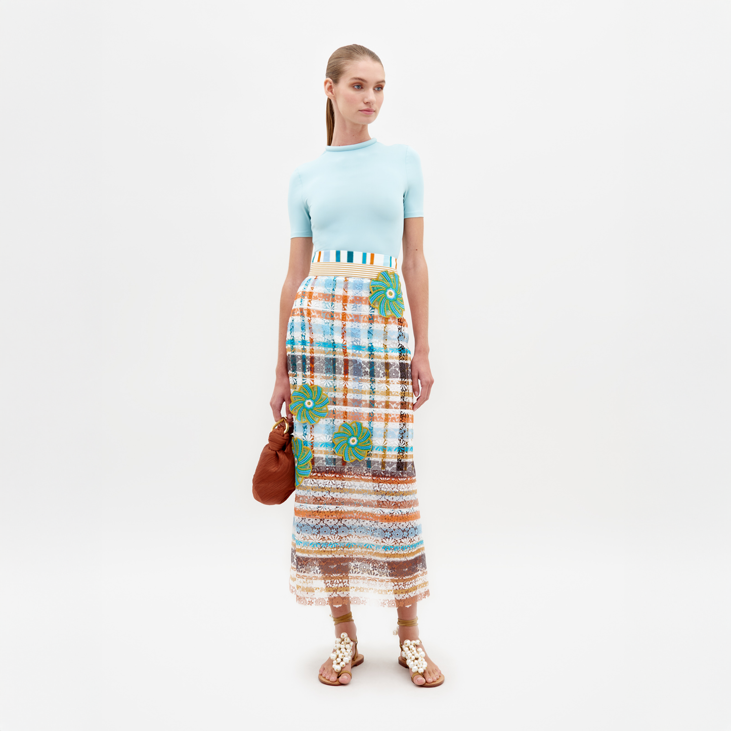 Against a white backdrop, a person models the Shadia Skirt by SVT DESIGNS—a high-waisted midi skirt with 3D floral appliqués—paired with a light blue top, while holding a brown purse and wearing embellished sandals.