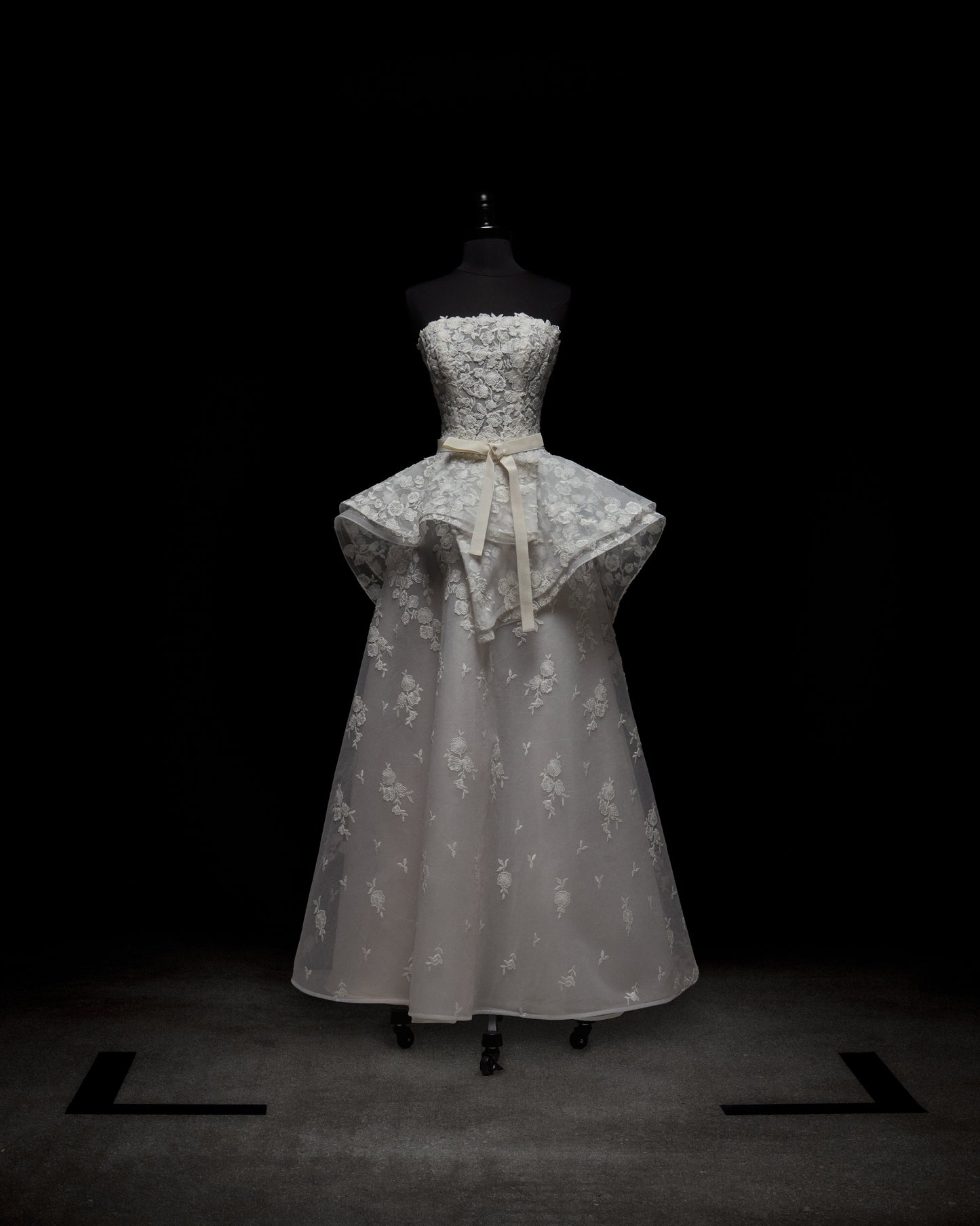 The Viviana Dress, a strapless white lace maxi with floral embroidery, fitted bodice, peplum overskirt, and ribbon belt, is displayed on a black mannequin against a dark background.