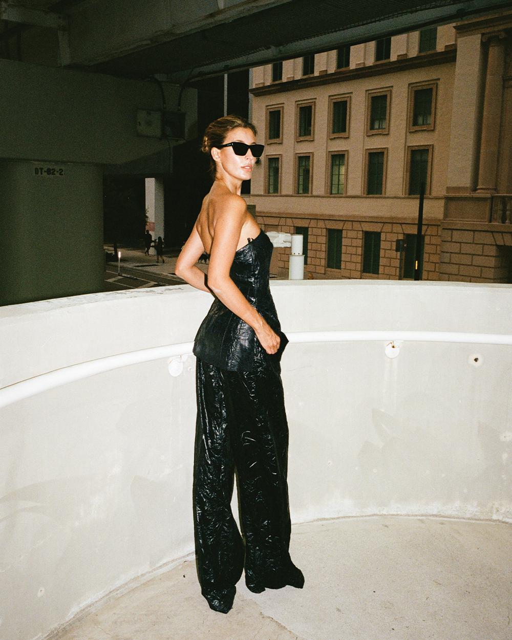 Woman in a black strapless top and pants standing on a rooftop with a cityscape background.