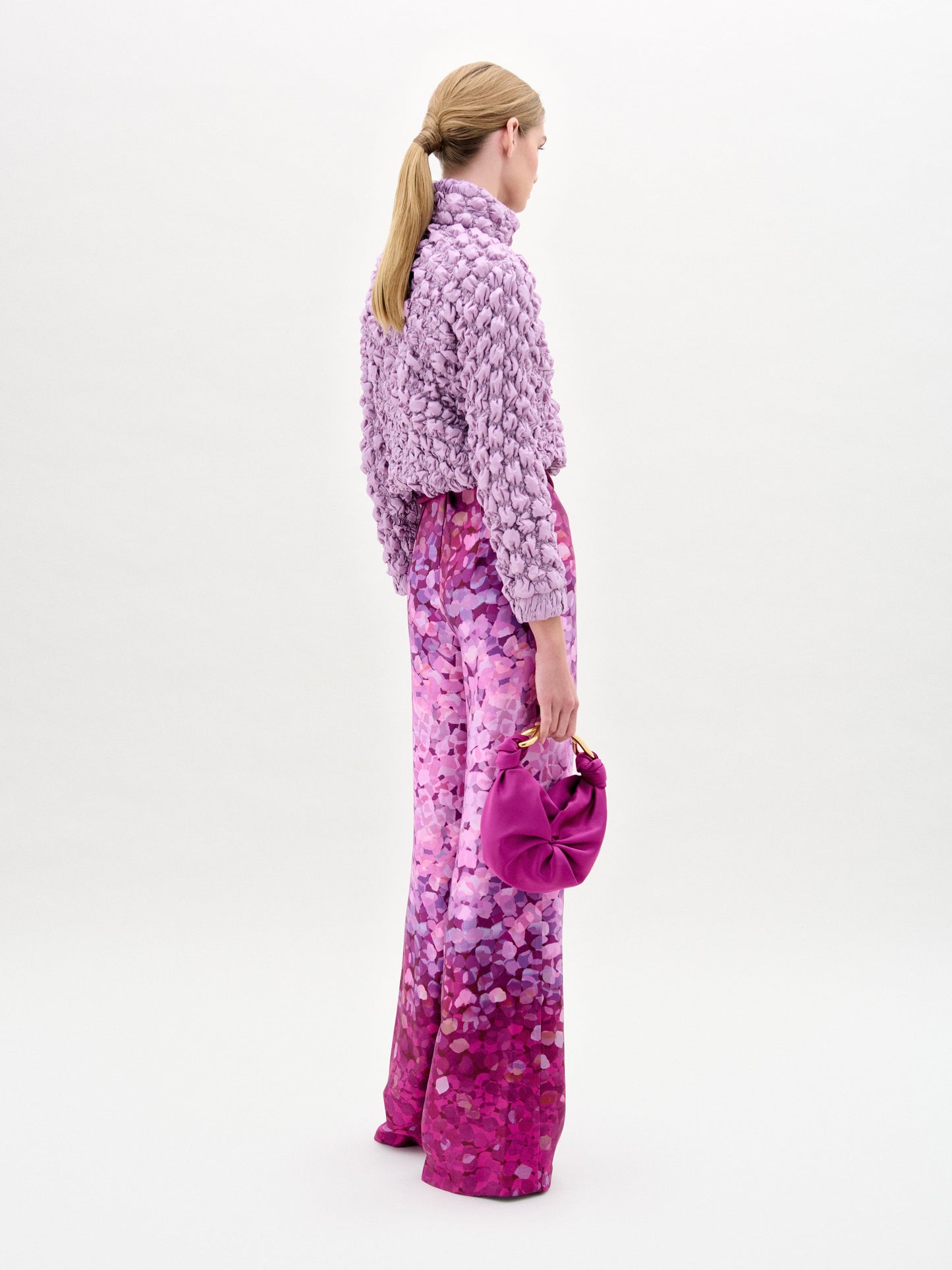 A woman poses against a white background wearing the oversized Zalta Jacket in textured lavender Italian fabric, paired with high-waisted pink patterned pants featuring a gold-buckled belt.