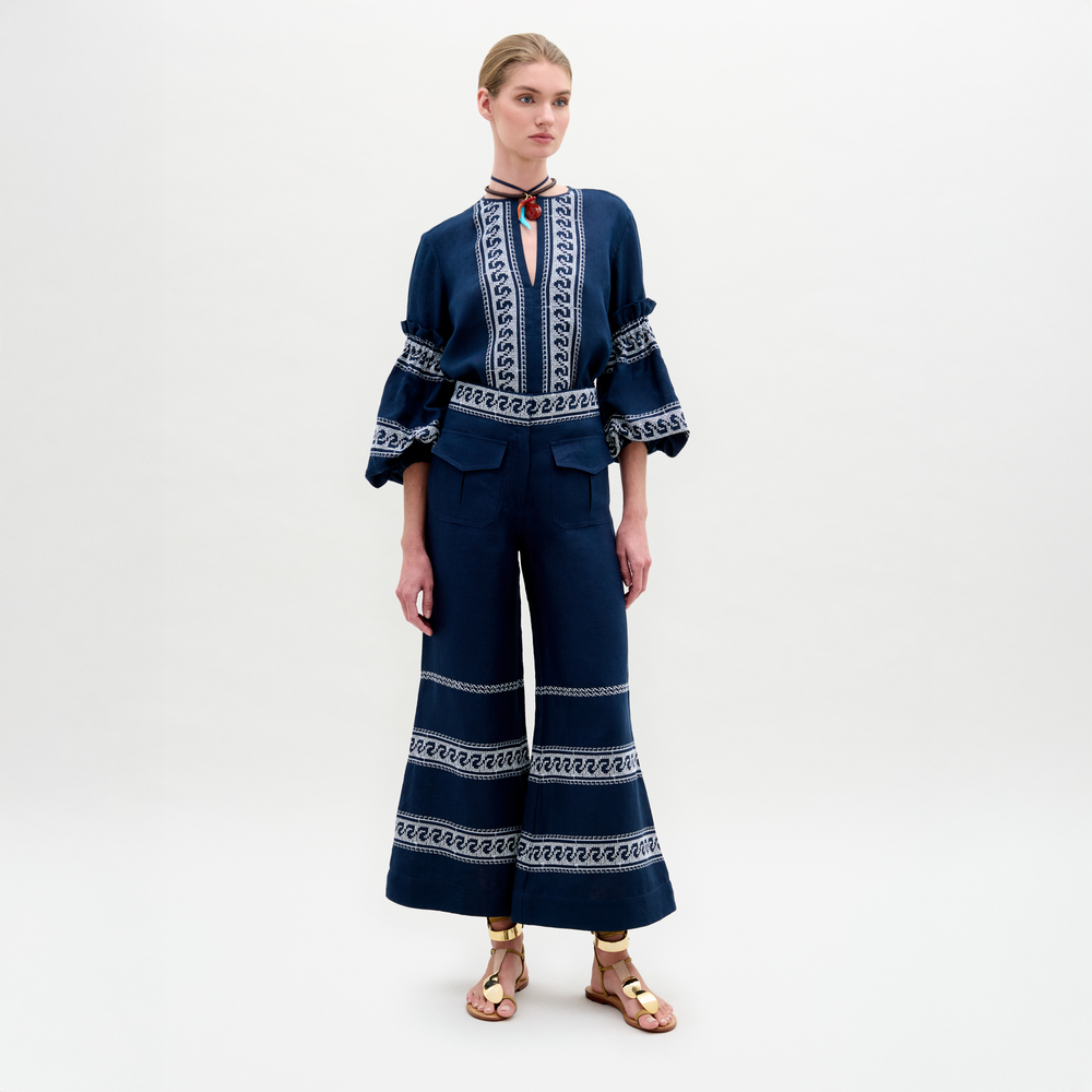 A woman poses against a plain backdrop wearing SVT DESIGNS’ Zina Pant—embroidered navy blue linen pants in a wide-leg silhouette—and a matching long-sleeve ruffled top, paired with gold sandals.