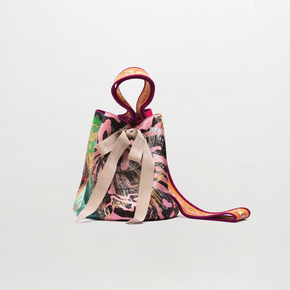 The Mochila Laaput Big by SVT DESIGNS is a multicolored hand-woven Wayuu handbag with pink, black, and green patterns, two vibrant handles, and a wide beige ribbon bow at the front, displayed against a plain light background.