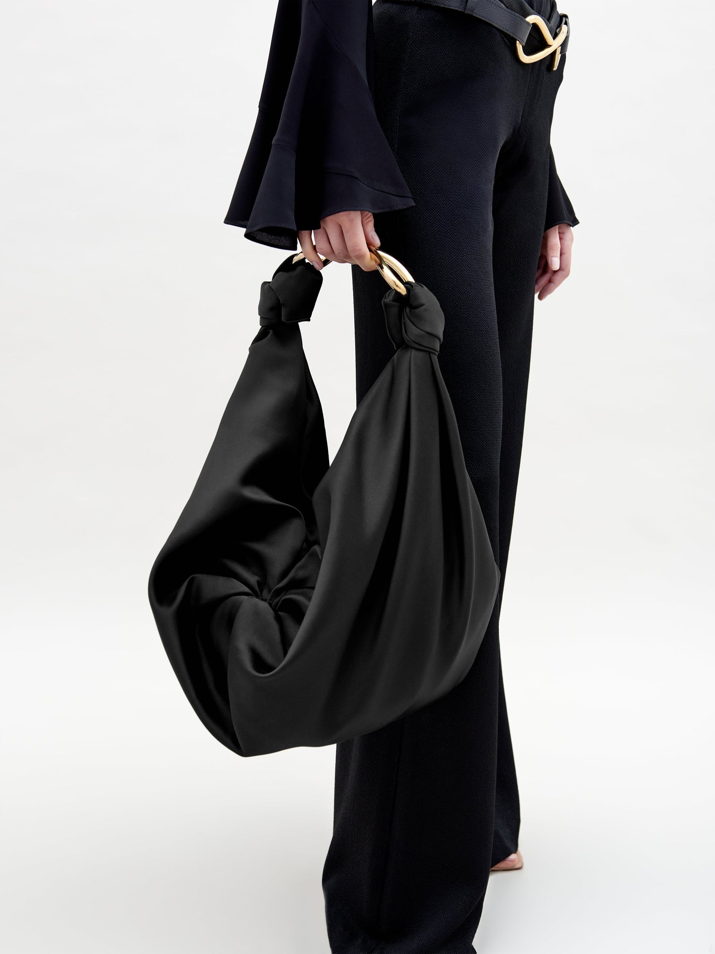 A person in black pants and a black top holds the Sofia Bag (Maxi), featuring gold handles and luxurious Italian textile, against a plain, light background that highlights the chic outfit and elegant accessory.