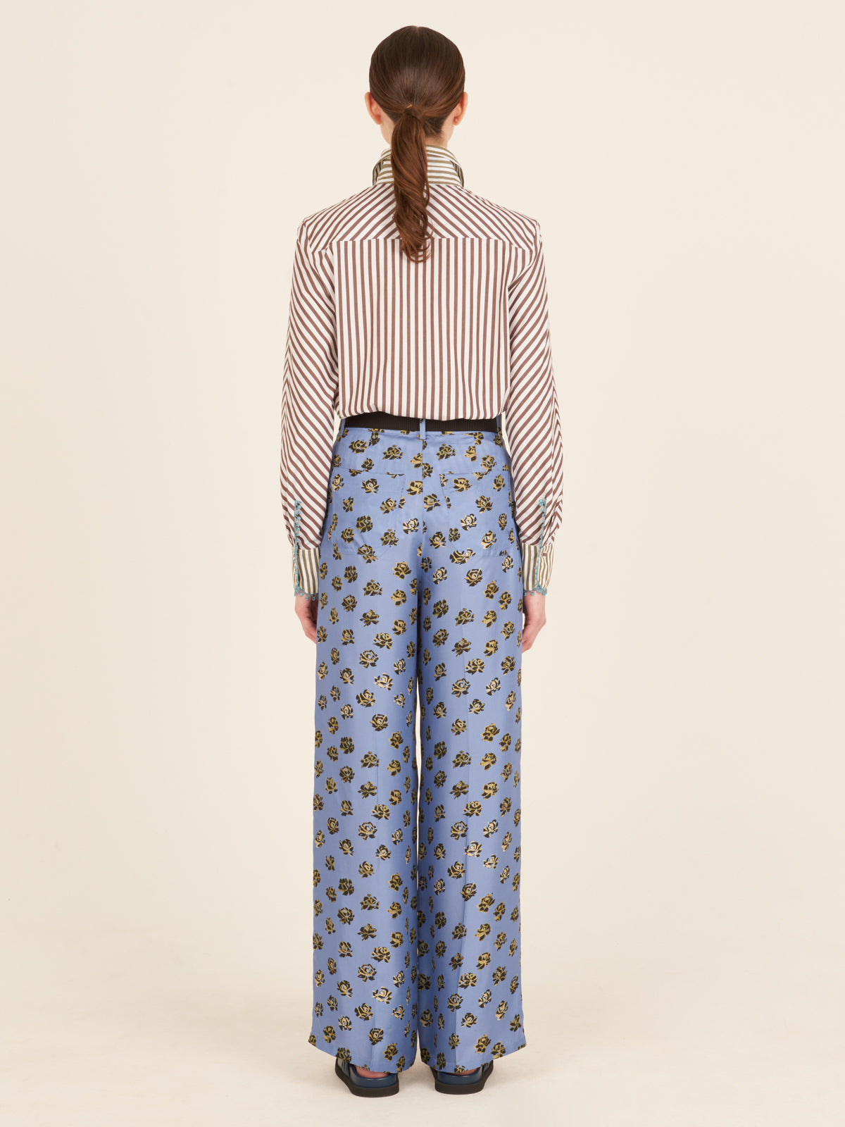 A person stands facing away from the camera, wearing the Silvia Tcherassi Melissa Blouse Brown Stripes tucked into high-waisted, wide-leg blue floral pants and paired with black shoes.