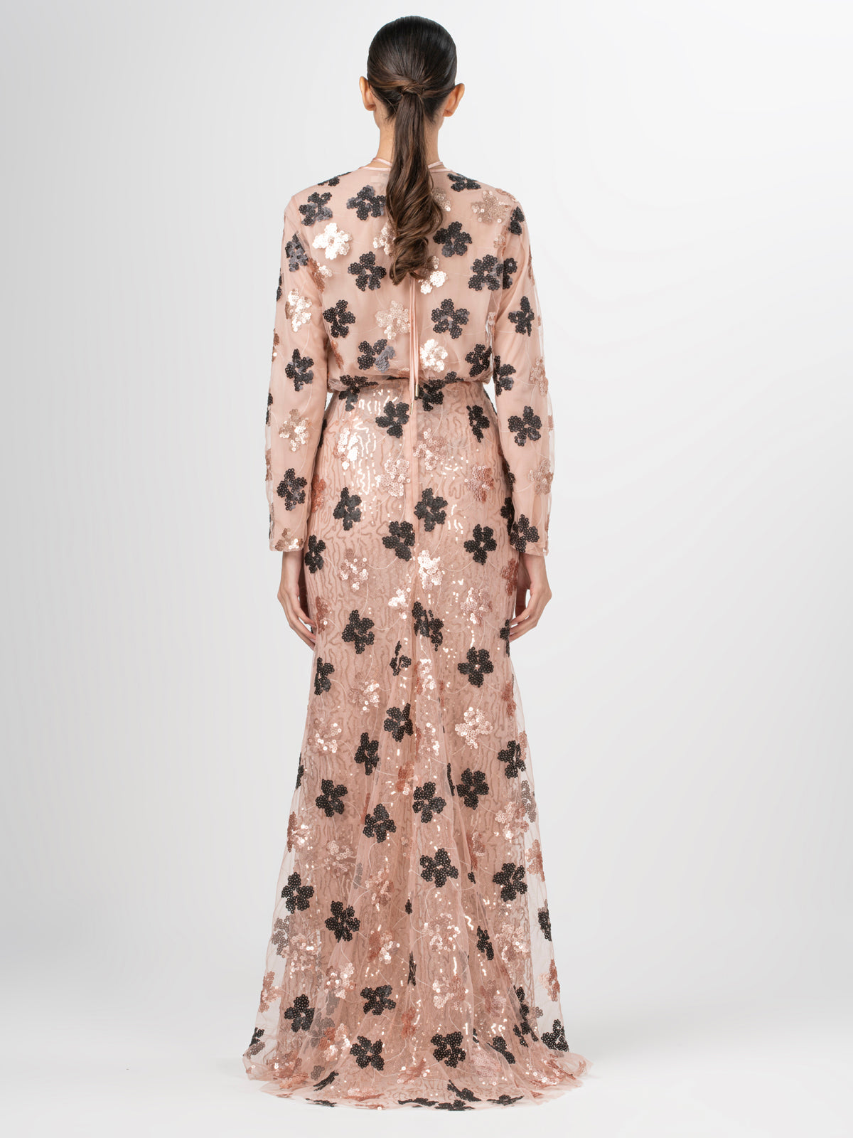 A person with long, dark hair in a ponytail models the Belisa Skirt from Silvia Tcherassi US. The floor-length, sheer skirt features black floral sequins lace over a beige underlayer. They stand against a white background, highlighting the back of the skirt.