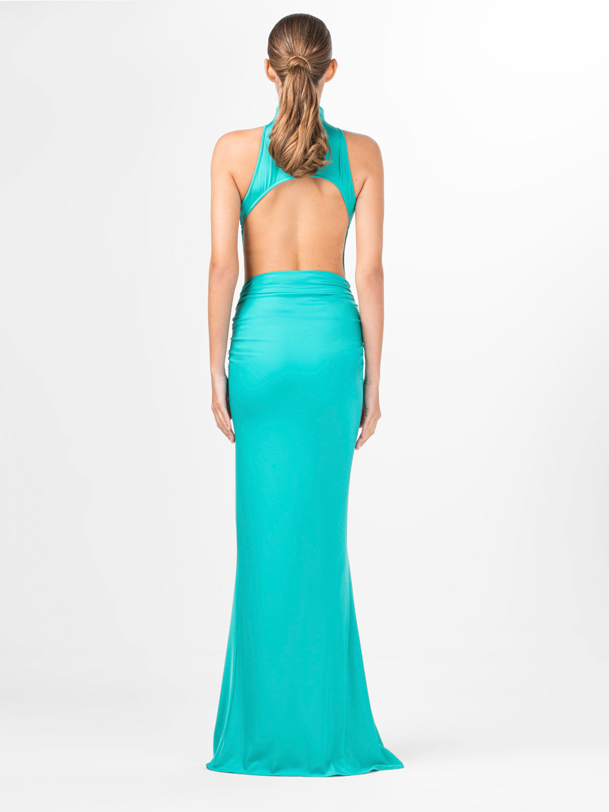 A woman with a brown ponytail stands with her back to the camera, wearing Sylvia Tcherassi US's turquoise Claudia Skirt. The long, backless skirt features an asymmetric waistline, halter neck, and draped silhouette against a plain white background.