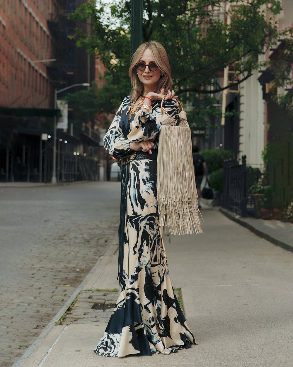 Silvia Tcherassi in a flowing, abstract-patterned dress and sunglasses stands confidently on a city sidewalk, holding the Eugenia Bag, with trees and buildings behind.