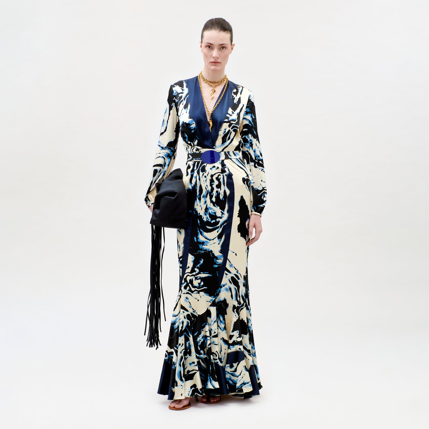 Model wearing a long, patterned dress with a black handbag on a white background