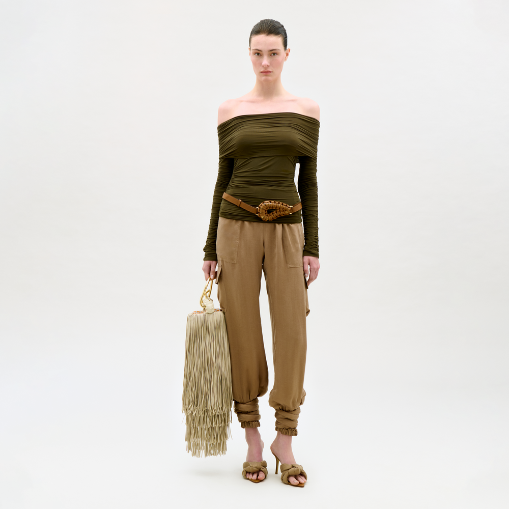 Woman wearing an off-shoulder green top and beige pants, holding a fringed beige bag on a white background