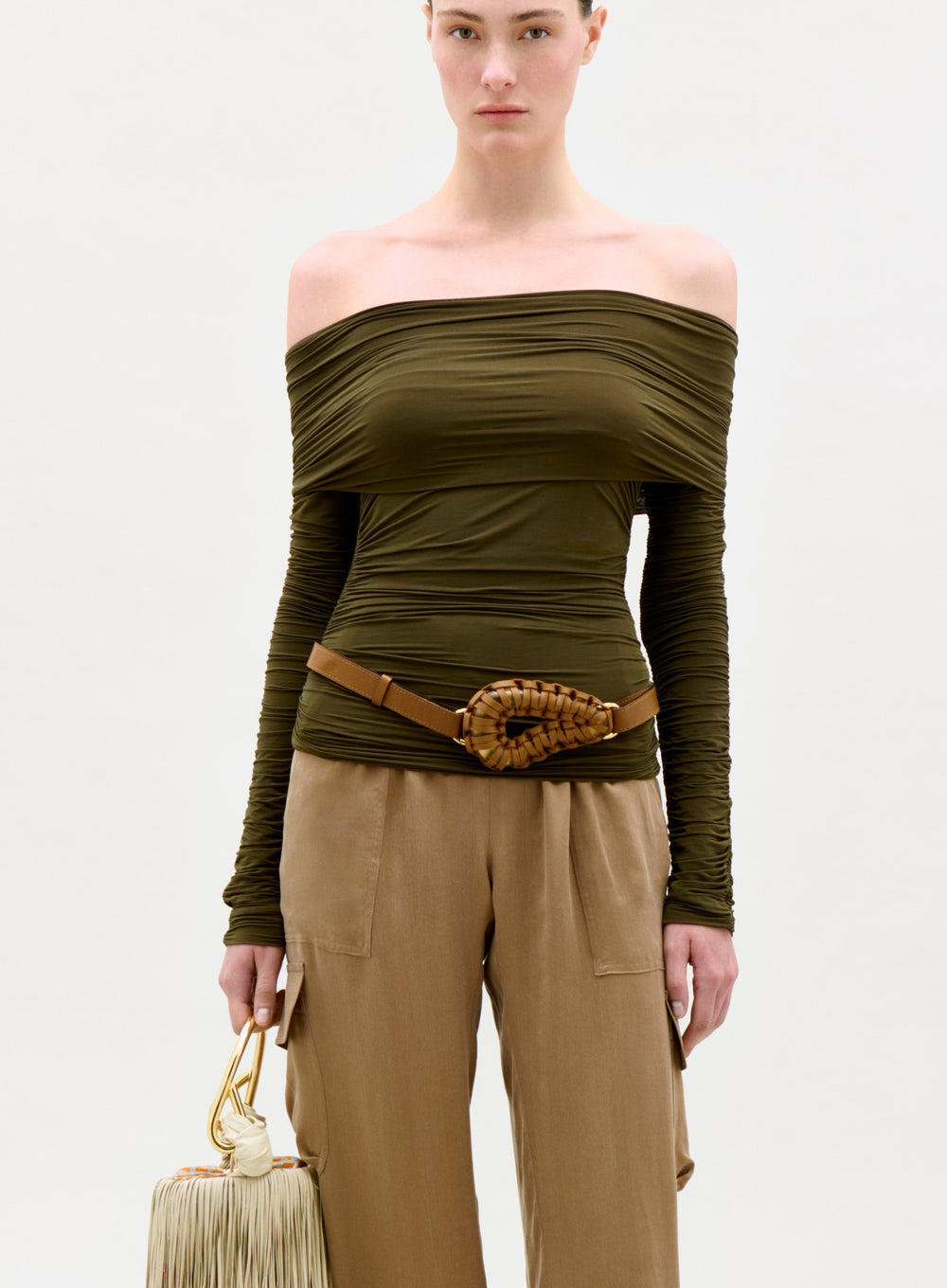 Woman wearing an olive green off-shoulder top and beige pants, holding a beige handbag on a white background.