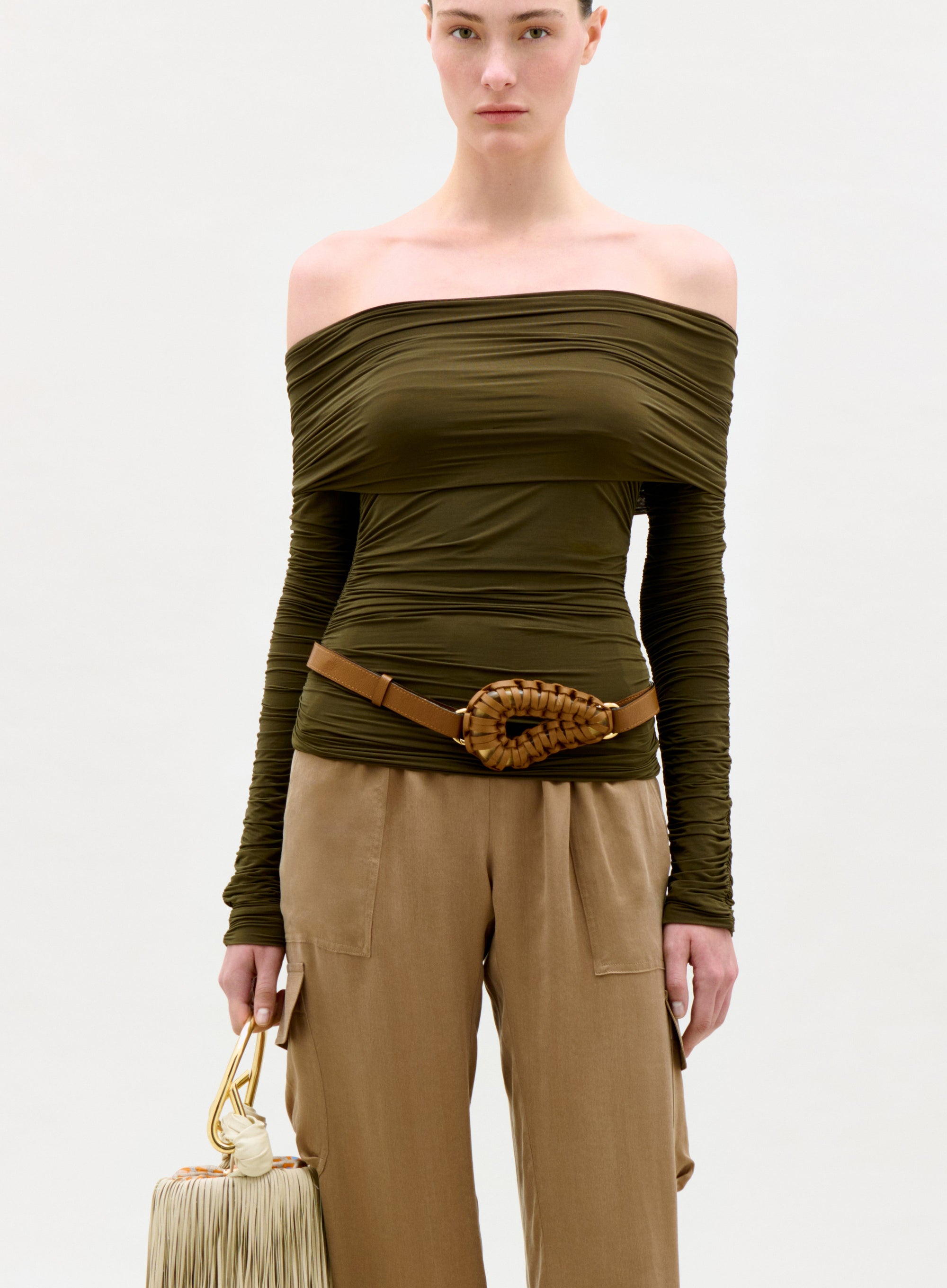 Woman wearing an olive green off-shoulder top and beige pants, holding a beige handbag on a white background.