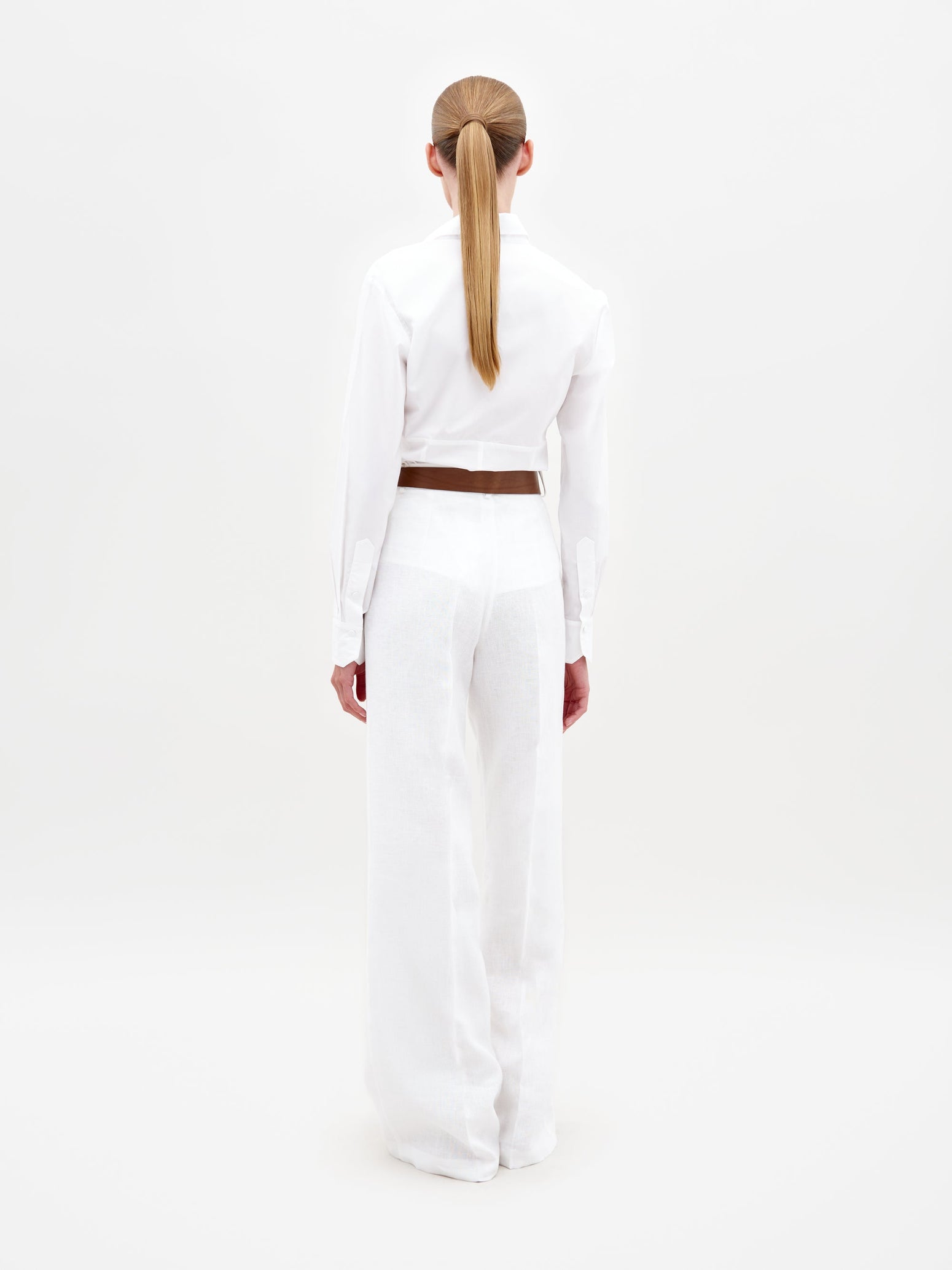 A woman is standing, wearing a white blouse with floral cut-out designs and high-waisted Trilce Pant White trousers. She has her hair pulled back and is wearing neutral-colored sandals. Pre-order by February 15th to ensure your outfit ships by date for your next event.