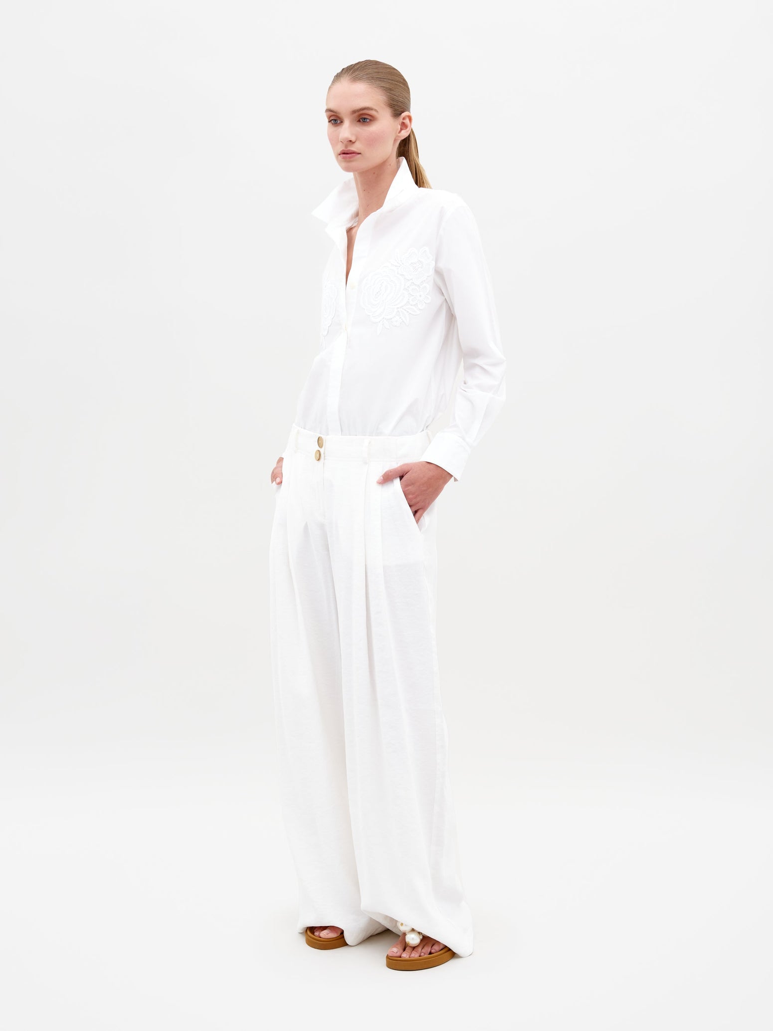 A person stands against a plain background, wearing an all-white outfit featuring the Opal Pant White—a pair of wide-legged pants—combined with a long-sleeve shirt with a high collar, their hands casually in their pockets. Pre-order now to secure your style for February 15th, 2025.