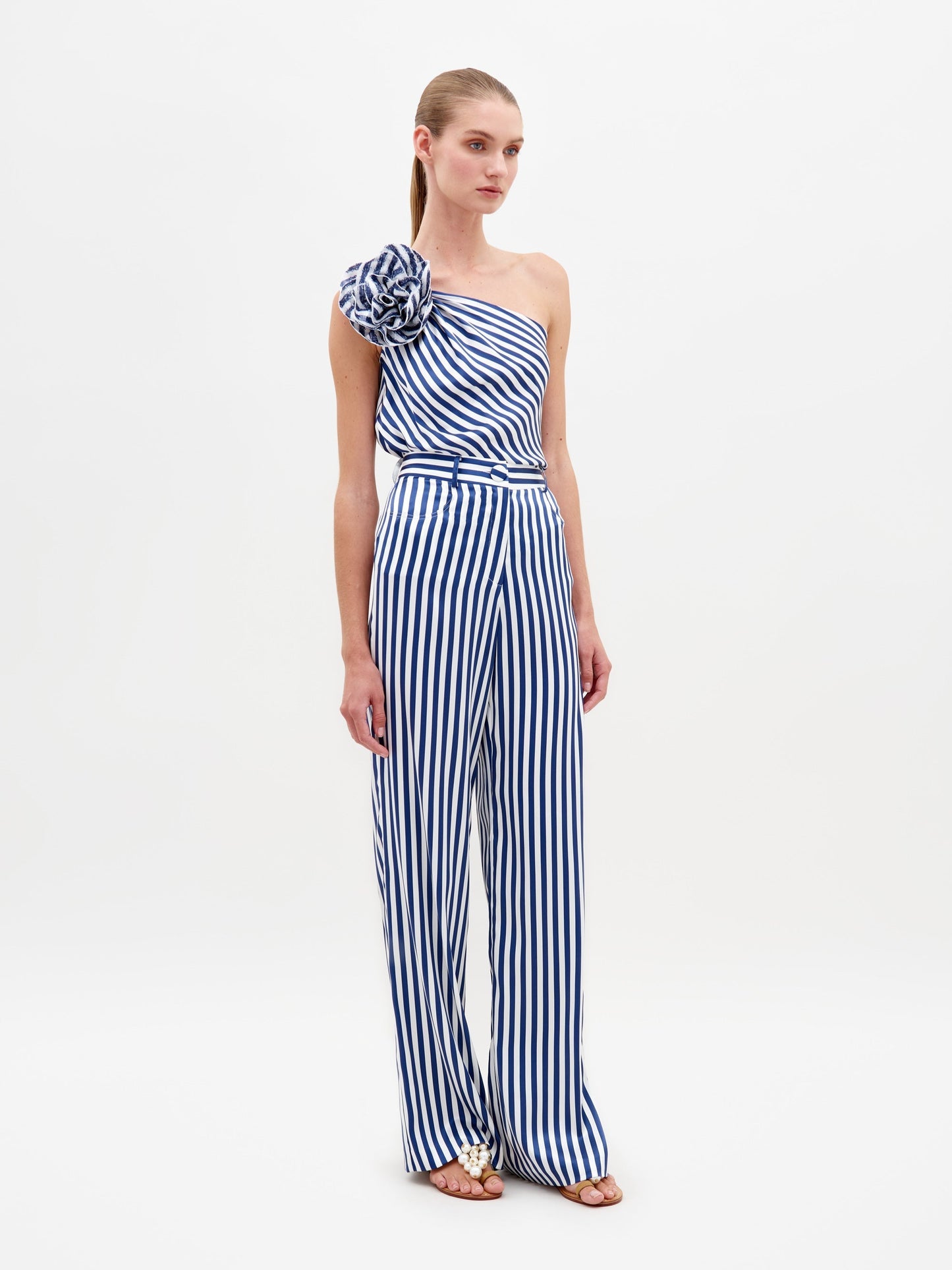 A woman stands wearing the Sebastiana Blouse in white and blue stripes, featuring a one-shoulder design with a large flower detail on the shoulder, against a plain white background. This chic outfit is available for pre-order now and will ship by February 15th, 2025.