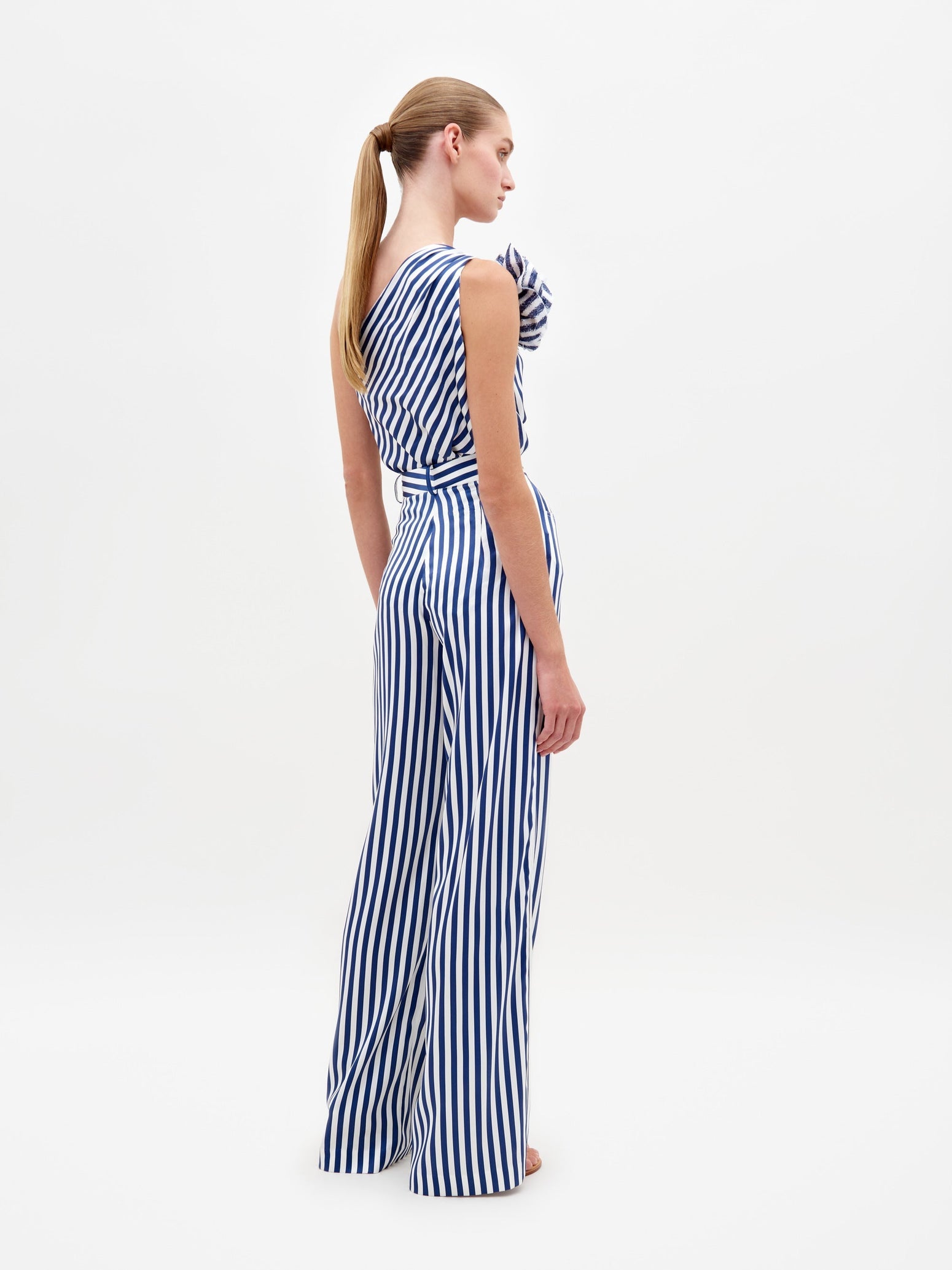 A woman stands wearing the Sebastiana Blouse in white and blue stripes, featuring a one-shoulder design with a large flower detail on the shoulder, against a plain white background. This chic outfit is available for pre-order now and will ship by February 15th, 2025.