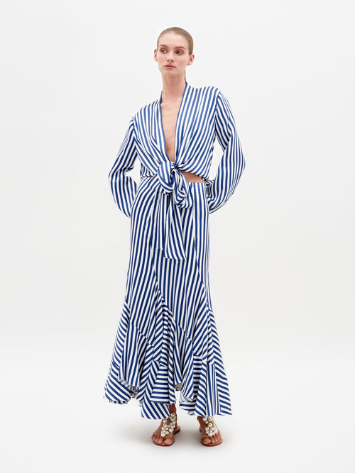 A person stands in the Honey Blouse White Blue Stripes, featuring a tie at the waist and a ruffled hem, paired with embellished sandals. This stylish ensemble is available for pre-order with shipping by February 15th.