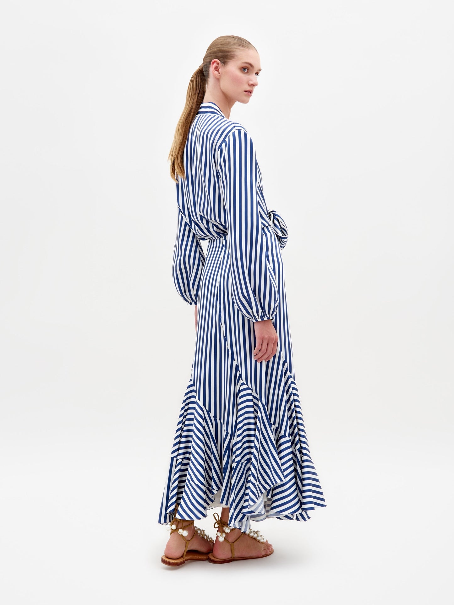 A person stands in the Honey Blouse White Blue Stripes, featuring a tie at the waist and a ruffled hem, paired with embellished sandals. This stylish ensemble is available for pre-order with shipping by February 15th.