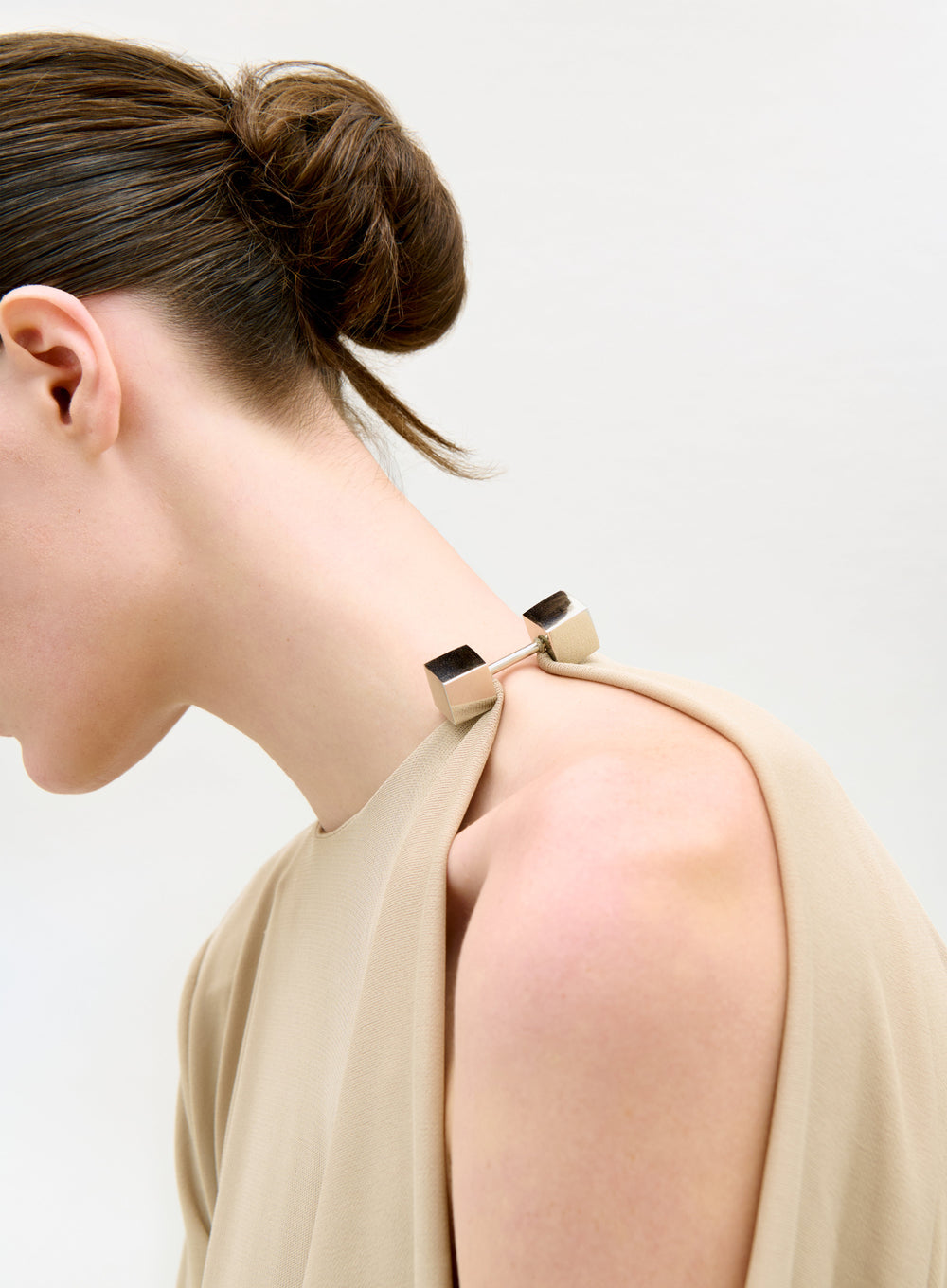 A person with dark hair in a sleek bun models the Natalia Dress—a beige, sleeveless piece fastened at the shoulder with a silver rectangular clasp—against a plain, light background.