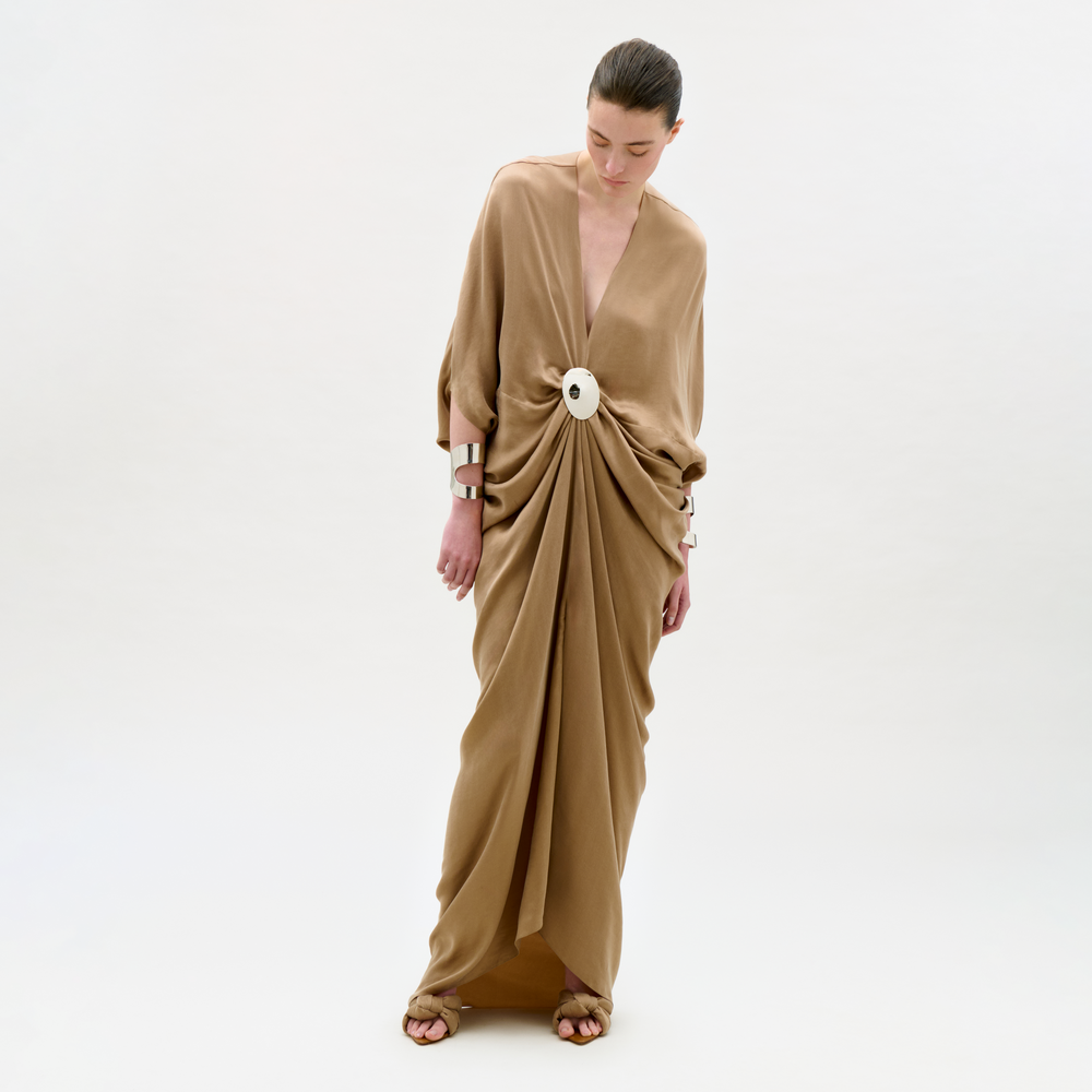 Woman wearing a long, flowing beige dress with a white belt on a plain background