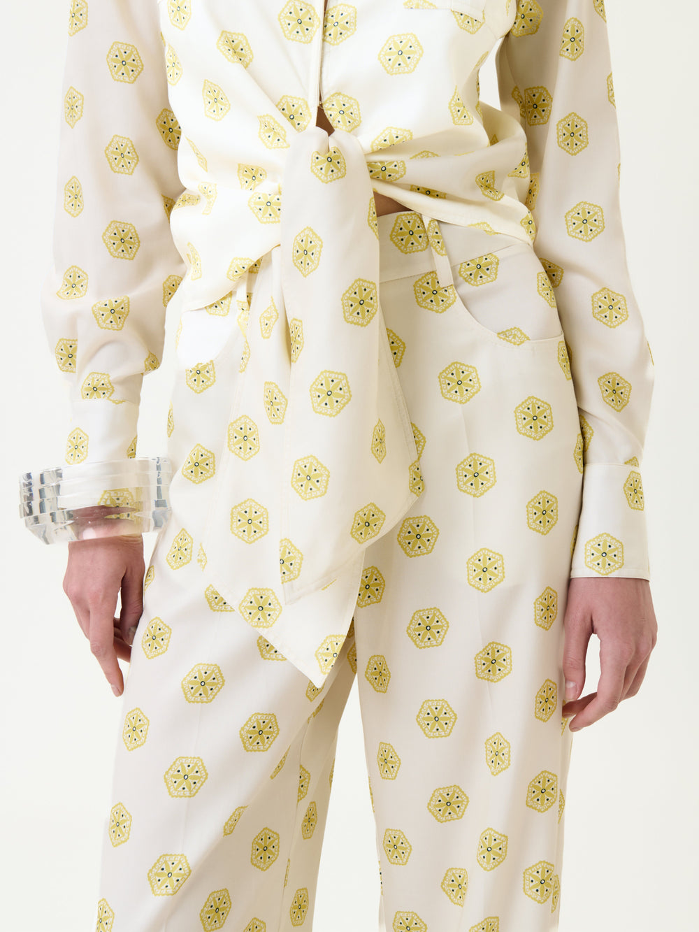 Model wearing the Caserta Blouse—a white top with a yellow geometric print, tied at the waist. Clear chunky bracelets adorn their wrist, and only the lower torso and upper legs are visible.