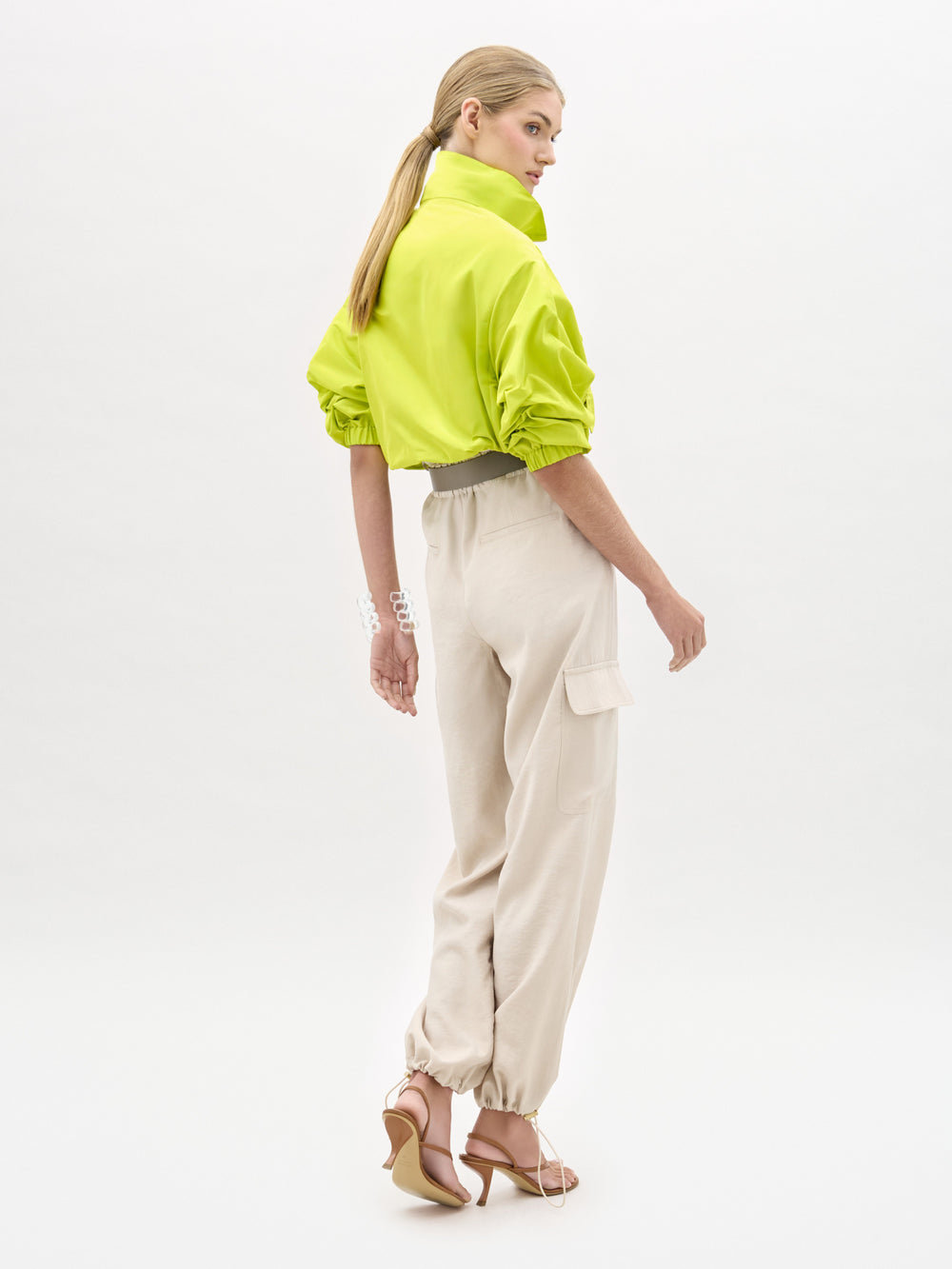 A person stands in profile against a white background wearing a neon yellow cropped jacket, SVT DESIGNS Darnell Pant in high-waist beige Italian fabric cargo style, and light-colored heeled sandals. Their hair is tied back in a low ponytail.