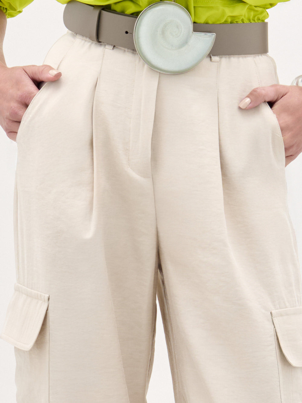 Wearing SVT DESIGNS’ Darnell Pant in beige high-waist Italian fabric with a wide belt and green spiral buckle, their hands are in the pockets, effortlessly paired with a bright green top.