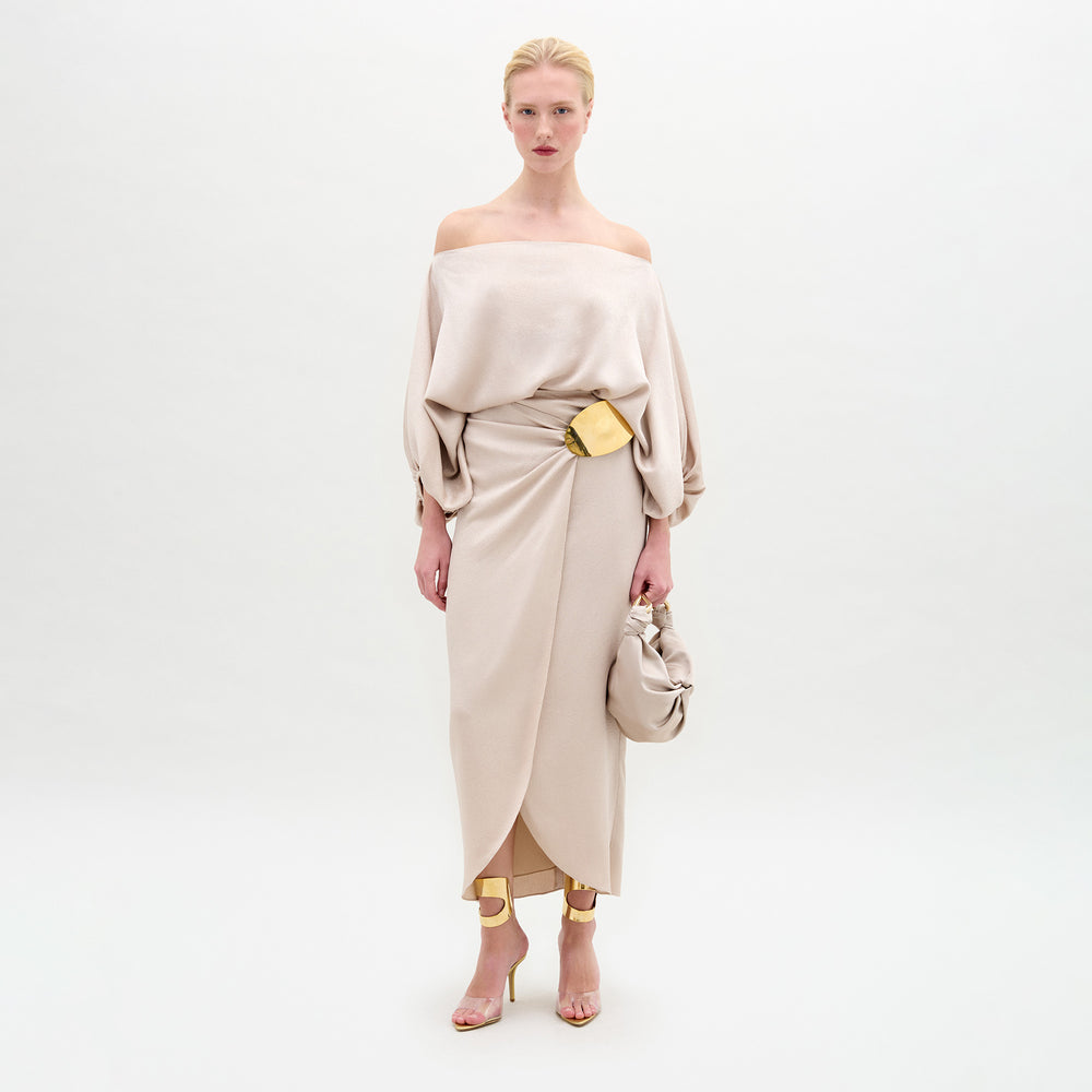A woman poses against a white background wearing the SVT DESIGNS Adrianne Skirt in beige hammered satin with draped sleeves, gold belt, gold ankle cuffs, high-heeled sandals, and a matching beige bag.