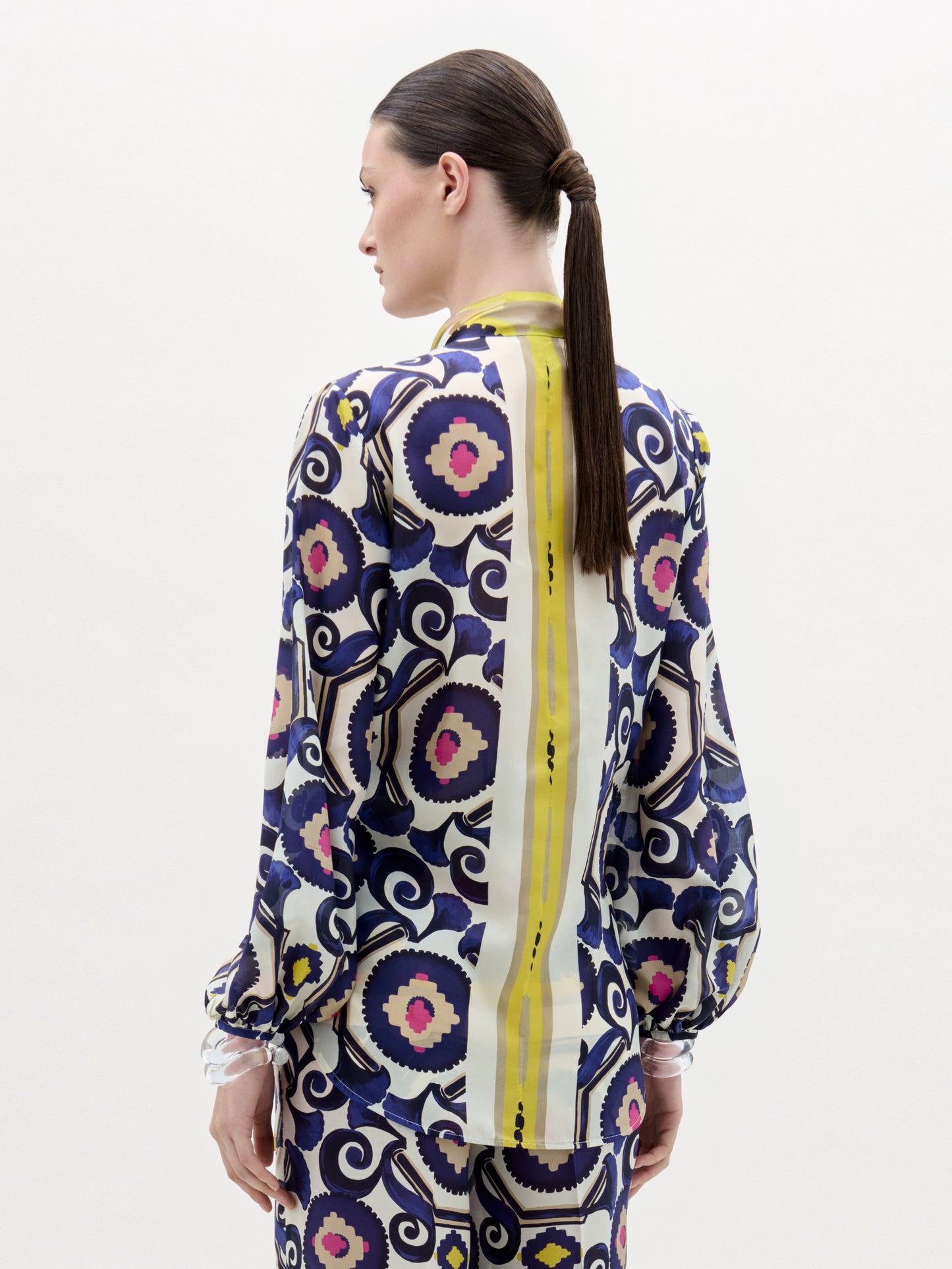 A person with straight brown hair in a low ponytail is shown from the back, wearing the SVT DESIGNS Honey Blouse—a long-sleeve silk top made from Italian fabric with bold geometric and floral prints and a vertical yellow stripe.
