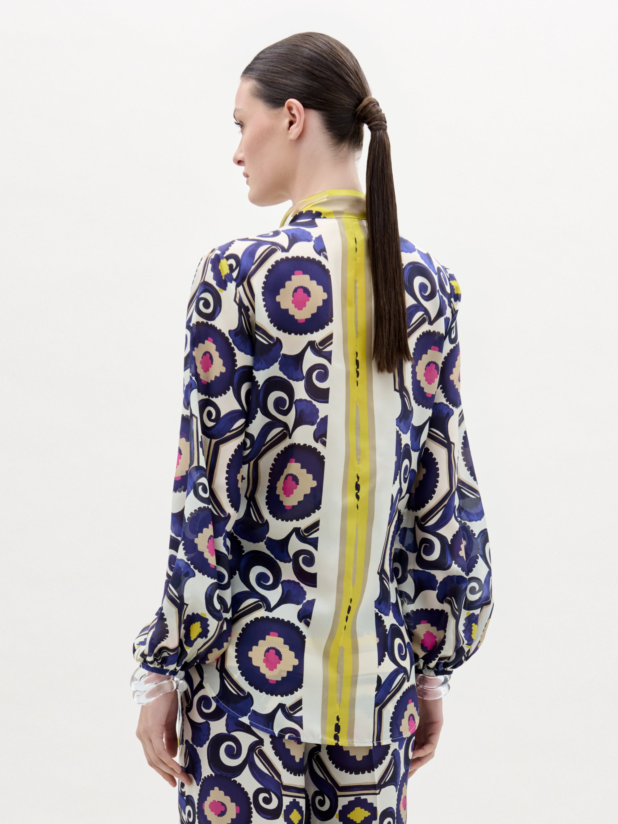 A person with straight brown hair in a low ponytail is shown from the back, wearing the SVT DESIGNS Honey Blouse—a long-sleeve silk top made from Italian fabric with bold geometric and floral prints and a vertical yellow stripe.