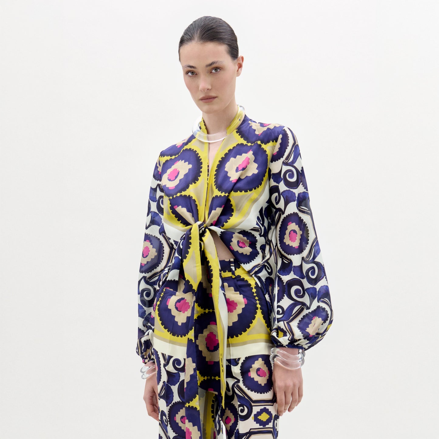 A woman stands against a plain white background wearing the SVT DESIGNS Honey Blouse—a silk top with yellow, blue, black, and pink circular and swirl patterns. Her hair is tied back and she has a neutral expression.