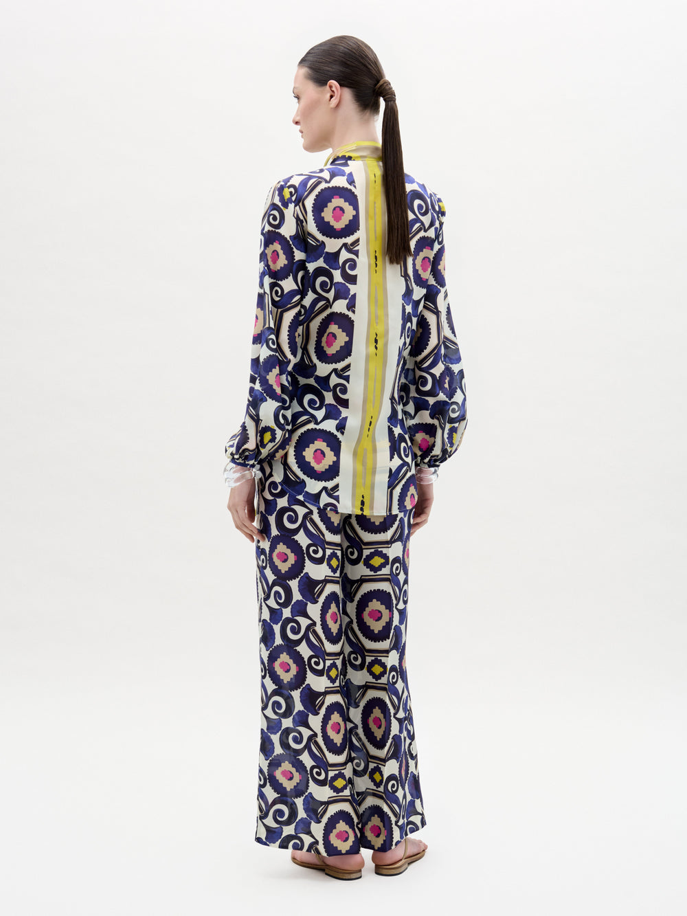 A person stands facing away, wearing the SVT DESIGNS Emine Pant—a long-sleeved, Italian-fabric outfit with purple, black, and beige circular patterns plus a bold vertical yellow stripe. Their hair is tied back in a low ponytail.