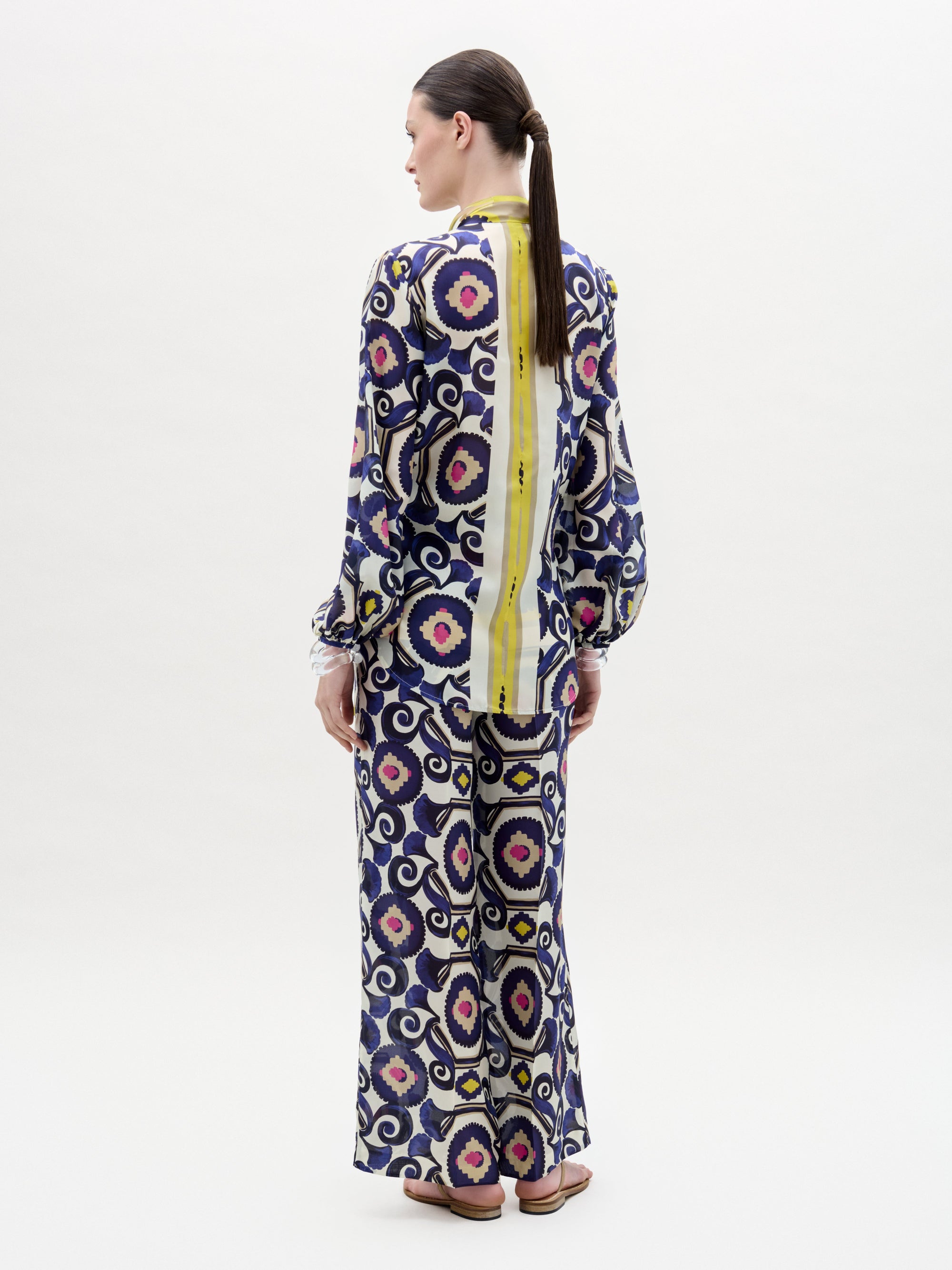 A person stands facing away, wearing the SVT DESIGNS Emine Pant—a long-sleeved, Italian-fabric outfit with purple, black, and beige circular patterns plus a bold vertical yellow stripe. Their hair is tied back in a low ponytail.