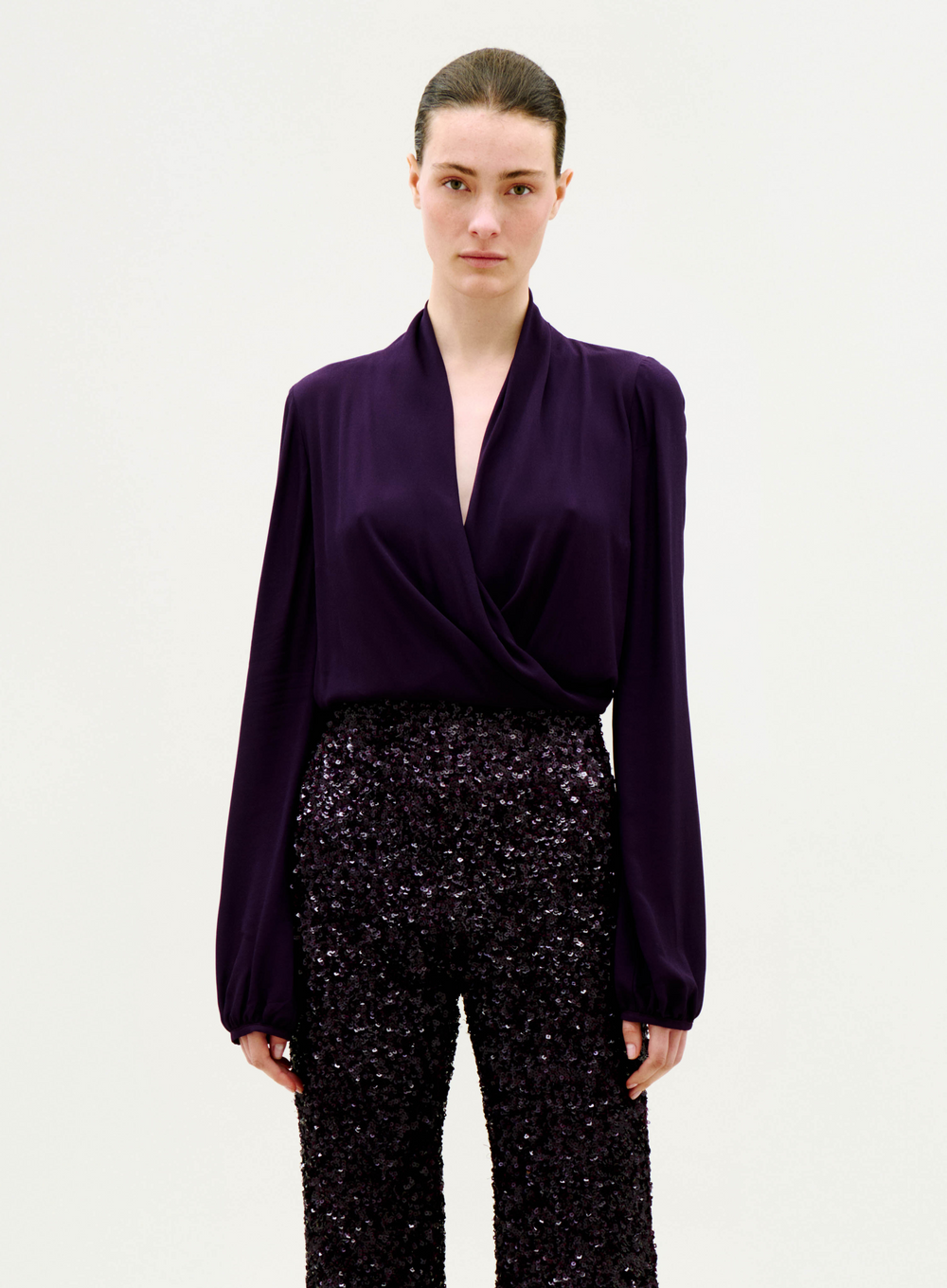 A woman with slicked-back hair wears the Tea Blouse—a deep purple, long-sleeve, wrap-style top—paired with high-waisted, dark sequin trousers, standing against a plain light background.