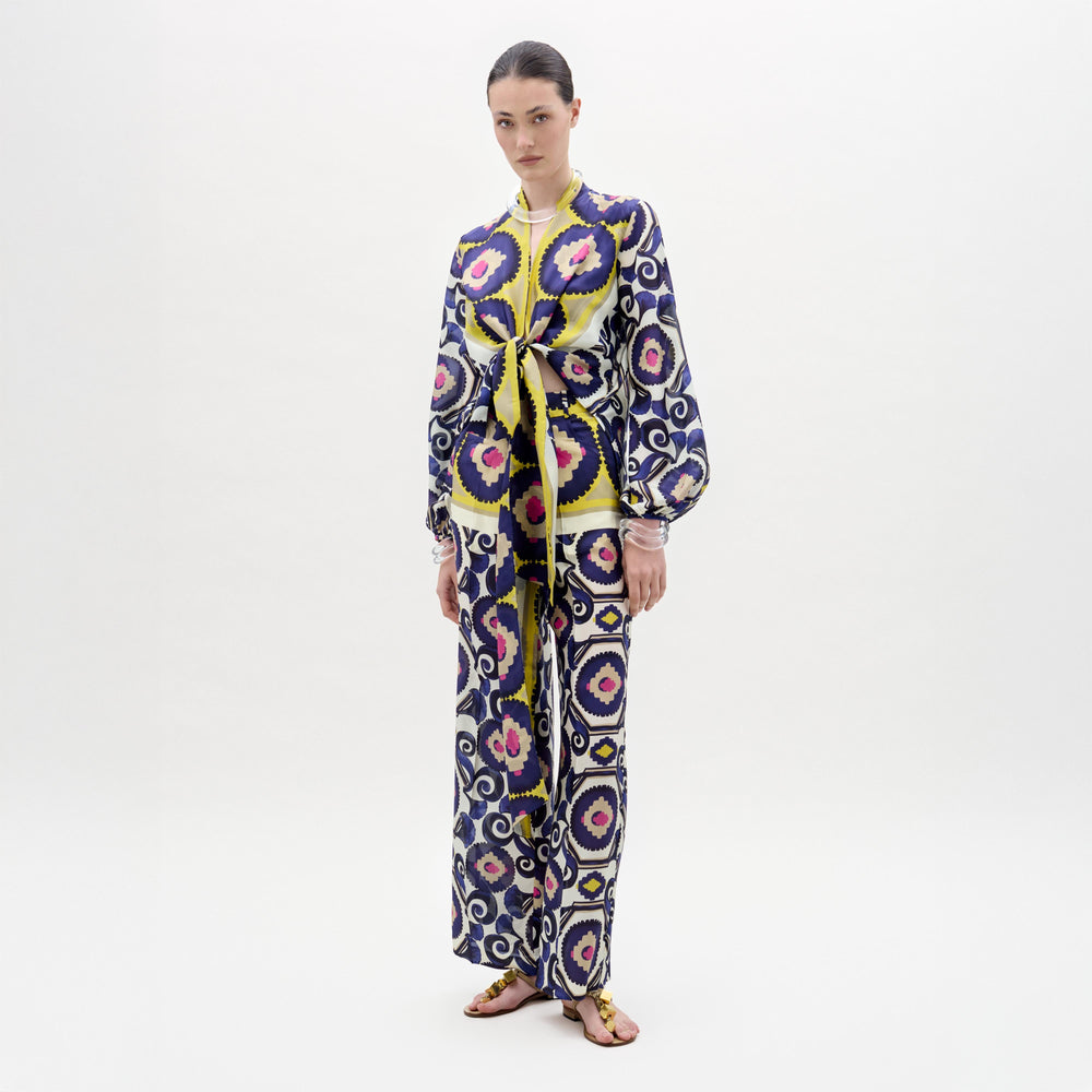 Against a plain white background, a woman models the Emine Pant by SVT DESIGNS, crafted from luxurious Italian fabric and featuring a bold geometric pattern in blue, yellow, pink, and white. She wears flat sandals with her hair pulled back.