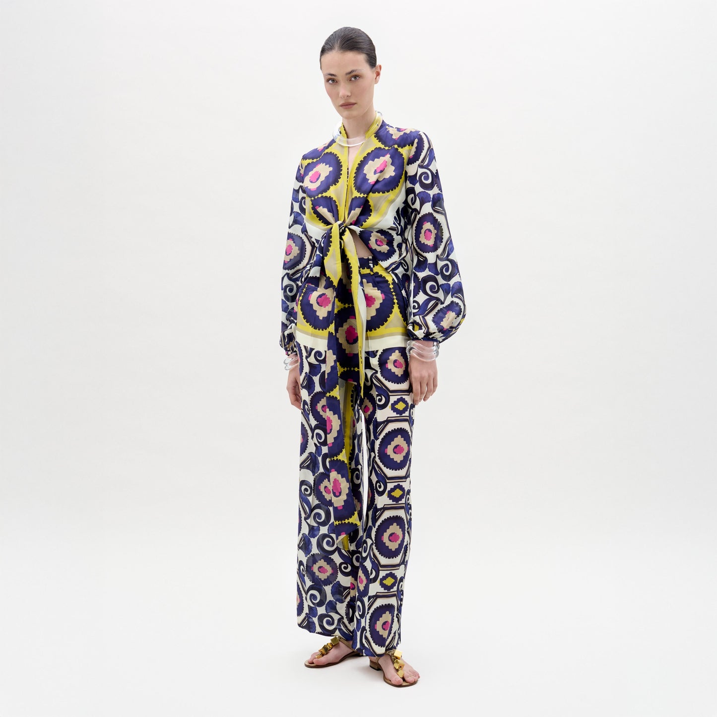 Against a plain white background, a woman models the Emine Pant by SVT DESIGNS, crafted from luxurious Italian fabric and featuring a bold geometric pattern in blue, yellow, pink, and white. She wears flat sandals with her hair pulled back.